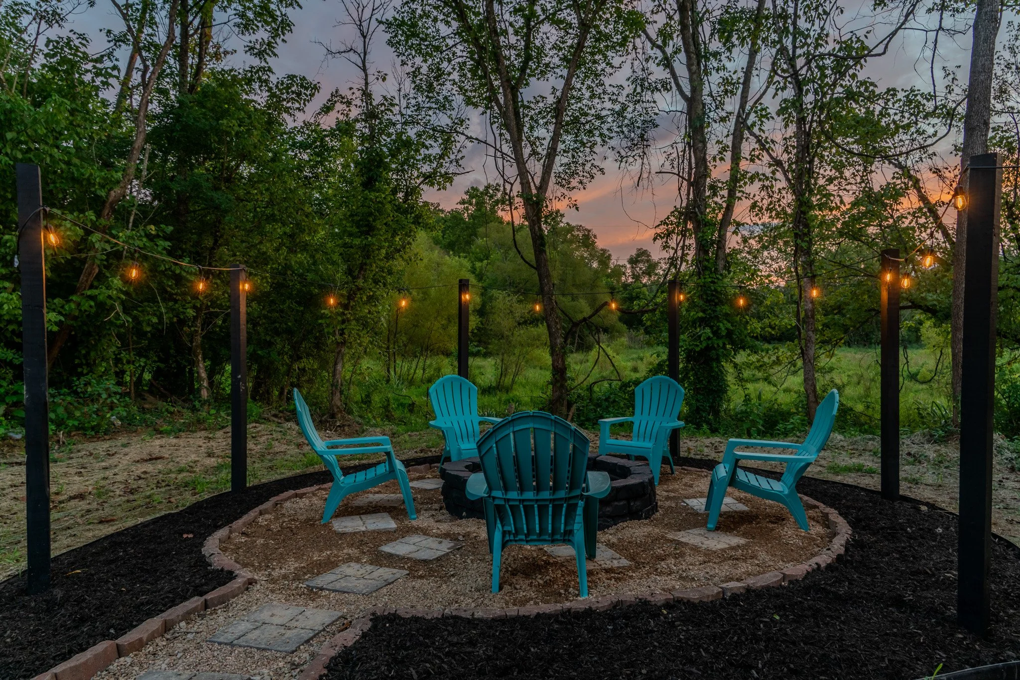 Five blue Adirondack chairs arranged in a circle around a fire pit in a backyard at dusk, with string lights hanging above and a wooded landscape in the background.