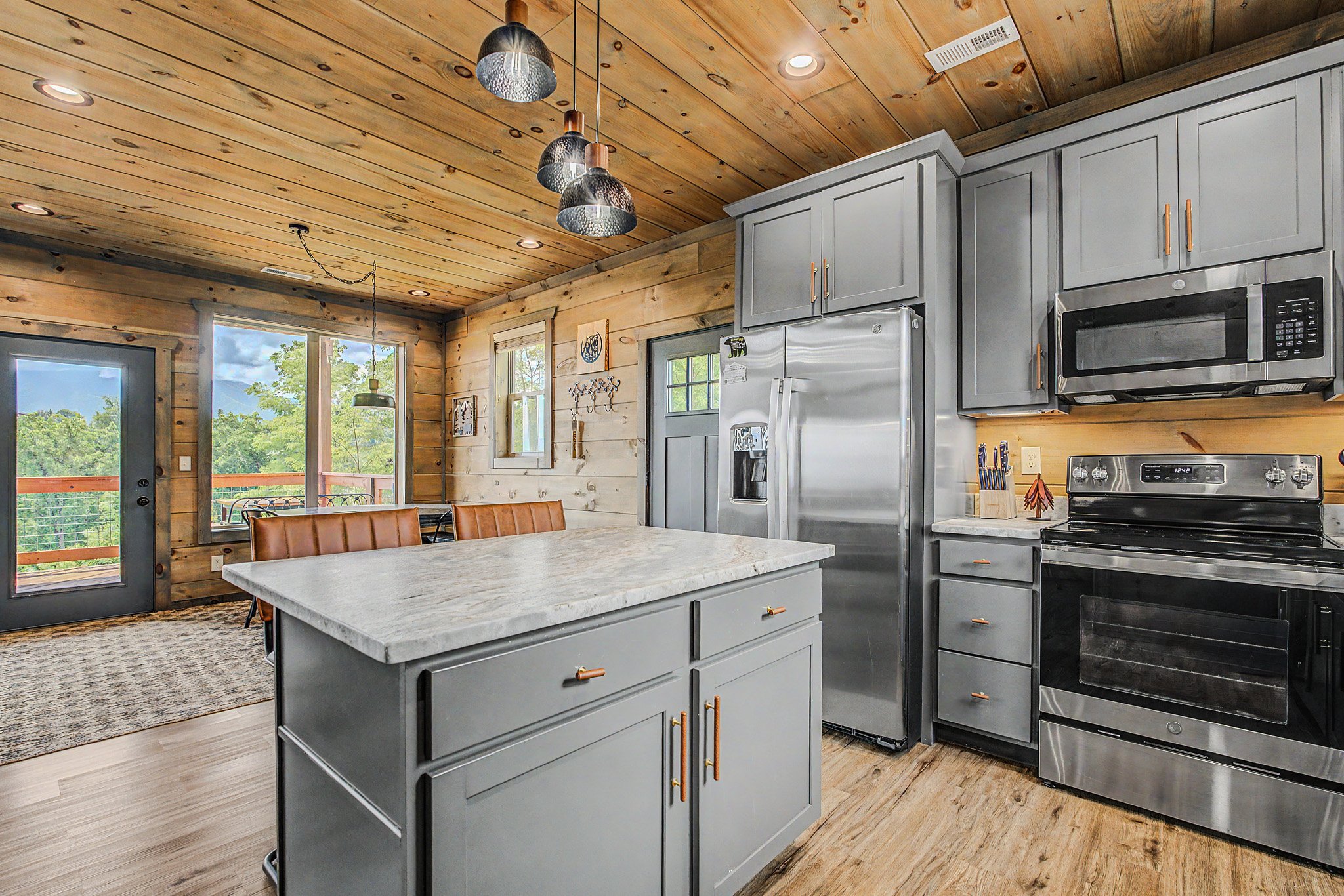 A rustic kitchen with wooden walls and ceiling, gray cabinets with gold handles, stainless steel appliances, and a kitchen island with a light-colored countertop, illuminated by pendant and recessed lighting.