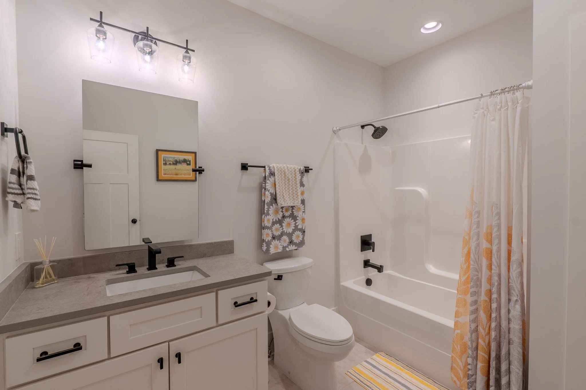 Bathroom with white walls, a vanity with a gray countertop, a mirror, black fixtures, a towel rack with a patterned towel, a toilet, and a bathtub with a shower curtain.