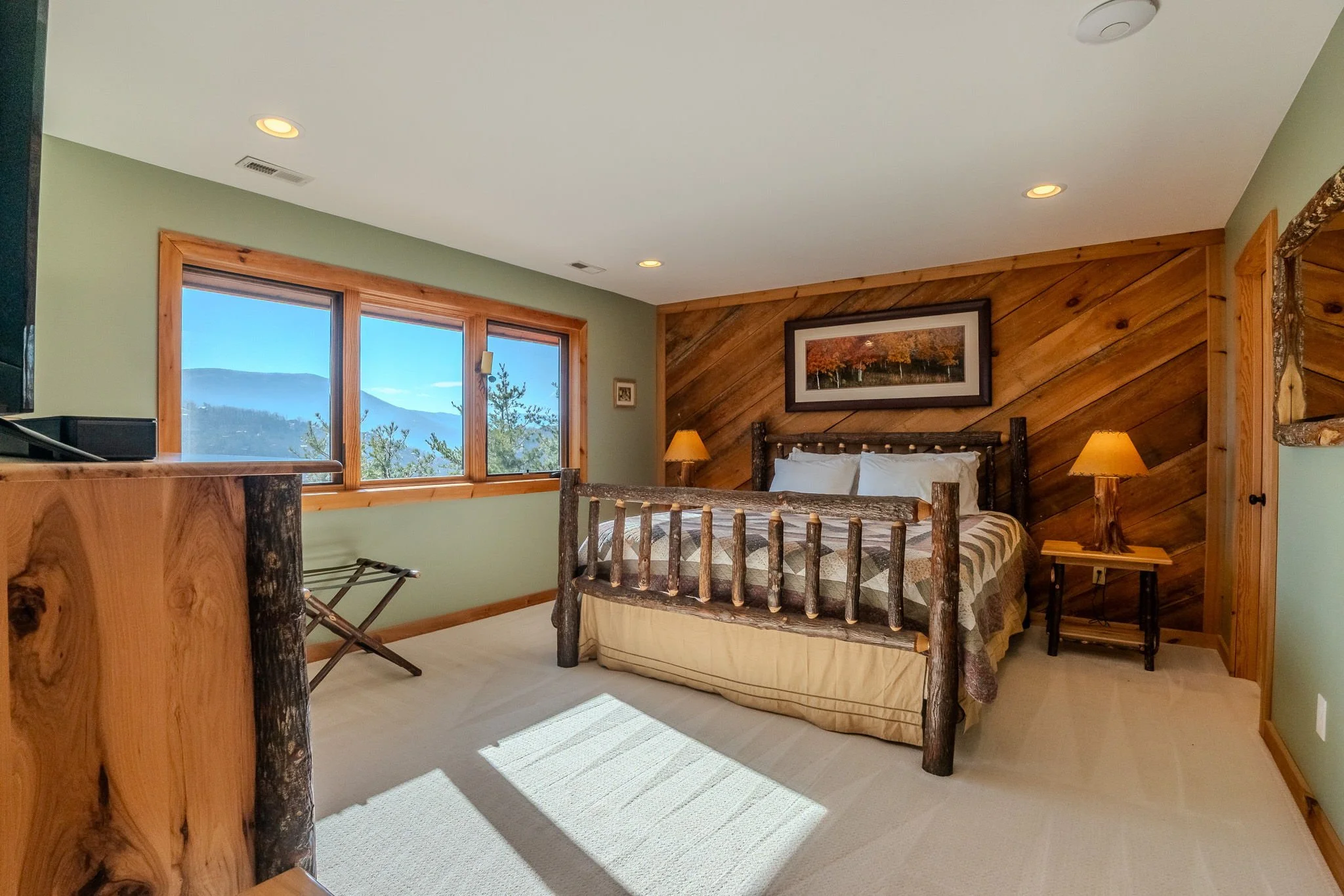 Cozy bedroom with wooden furniture, a large bed with pillows, a wooden wall with artwork, a window showing trees and mountains, and warm lighting.