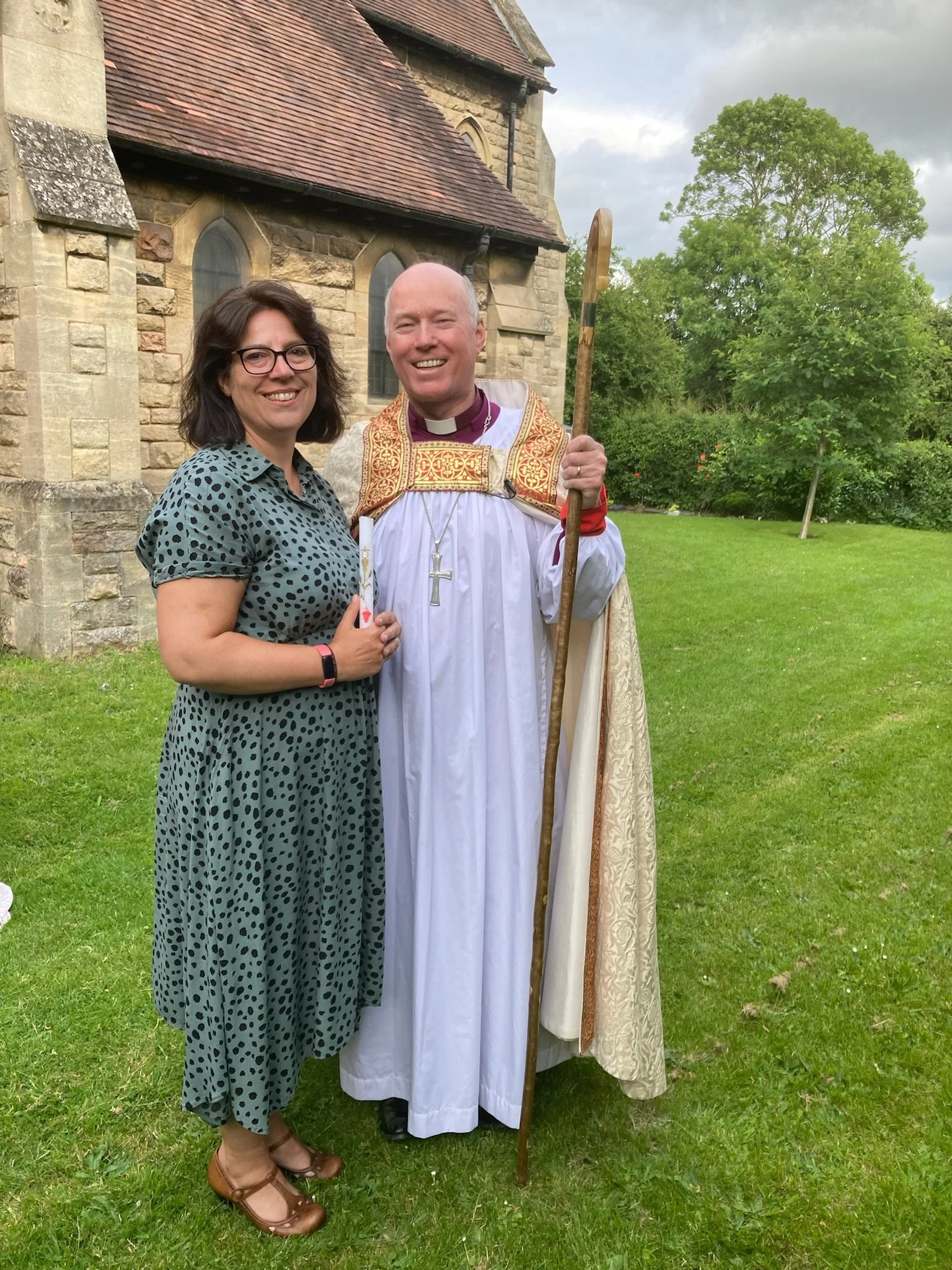 A woman and a priest standing outdoors on a grassy area in front of a stone church, smiling for the camera. The woman wears a polka-dot dress and glasses, while the priest is dressed in white ecclesiastical robes with a gold-embroidered cape, holding a wooden staff, and wearing a cross necklace.