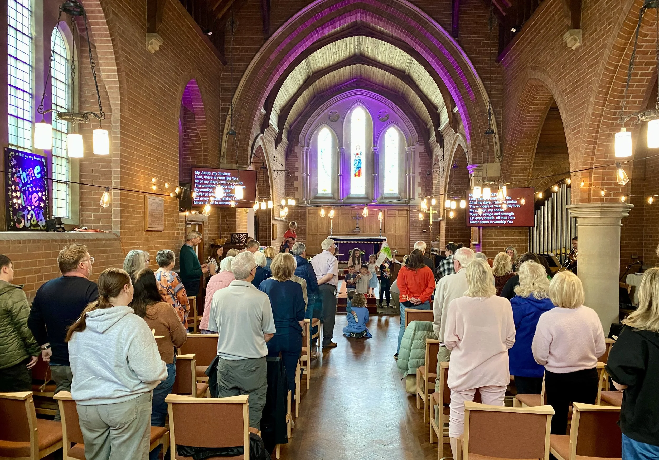 Congregation gathered in a church with brick walls, stained glass windows, and an altar, during a worship service or prayer gathering with worship screens showing lyrics.