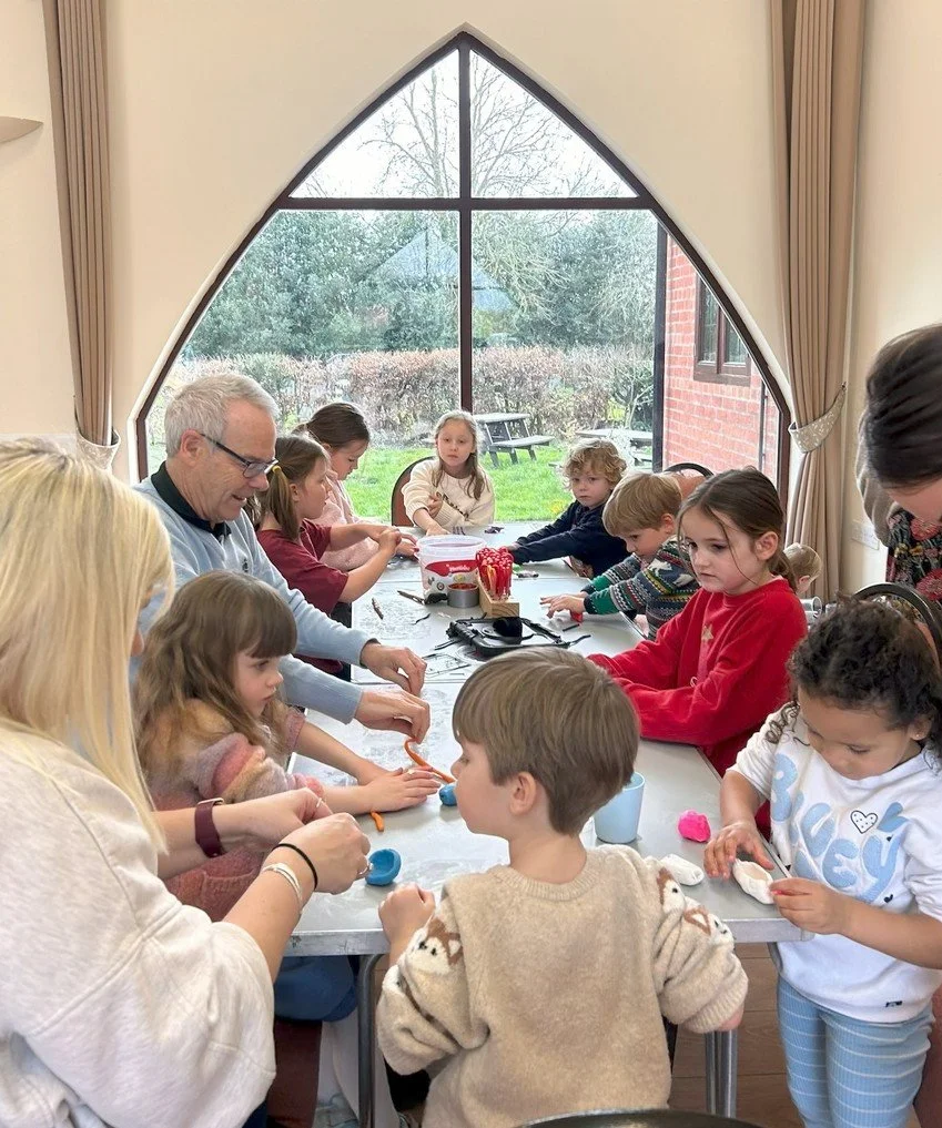 Children and adults sitting around a long table engaging in arts and crafts indoors, with a large arched window showing outdoor trees and a picnic table.