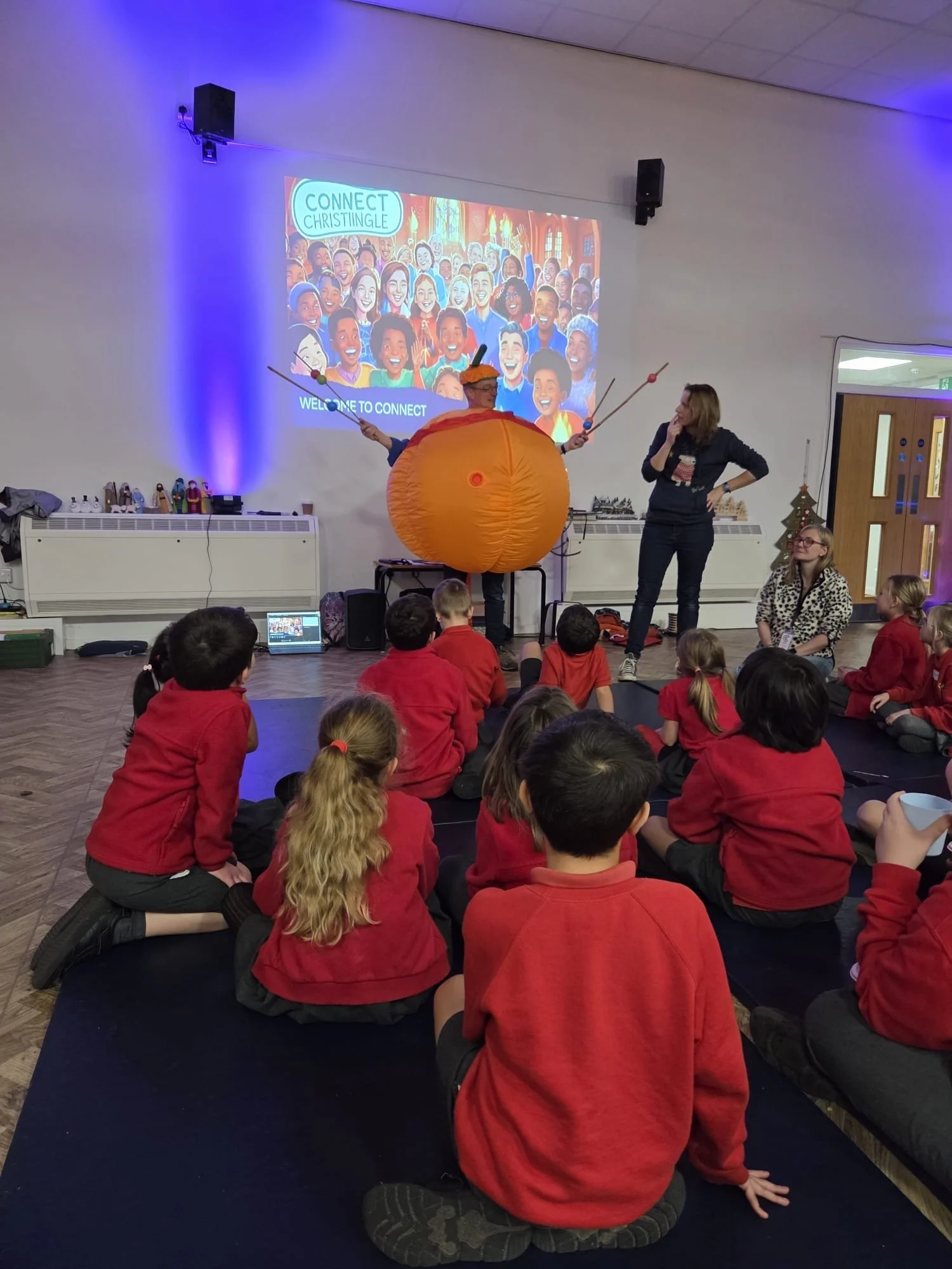Children sitting on the floor watching a performance in a classroom with a woman in a costume resembling a planet, with a large orange sphere and antennae, and a woman standing beside them. A colorful cartoon projection on the wall reads "Connect Christingle" and "Welcome to Connect."