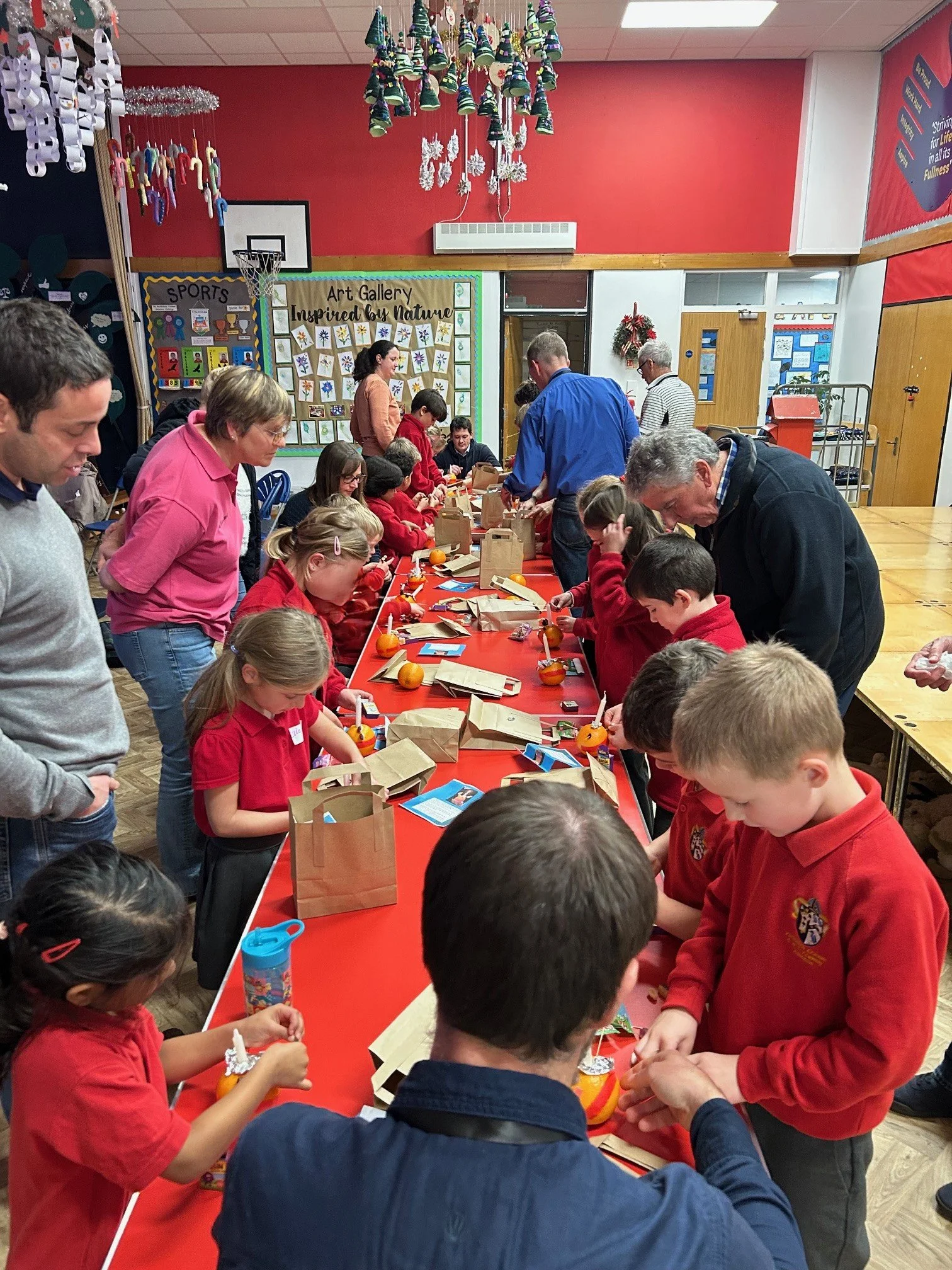 Children and adults gather around a long red table, engaged in craft activities, in a school or community center decorated with Christmas ornaments and holiday artwork.