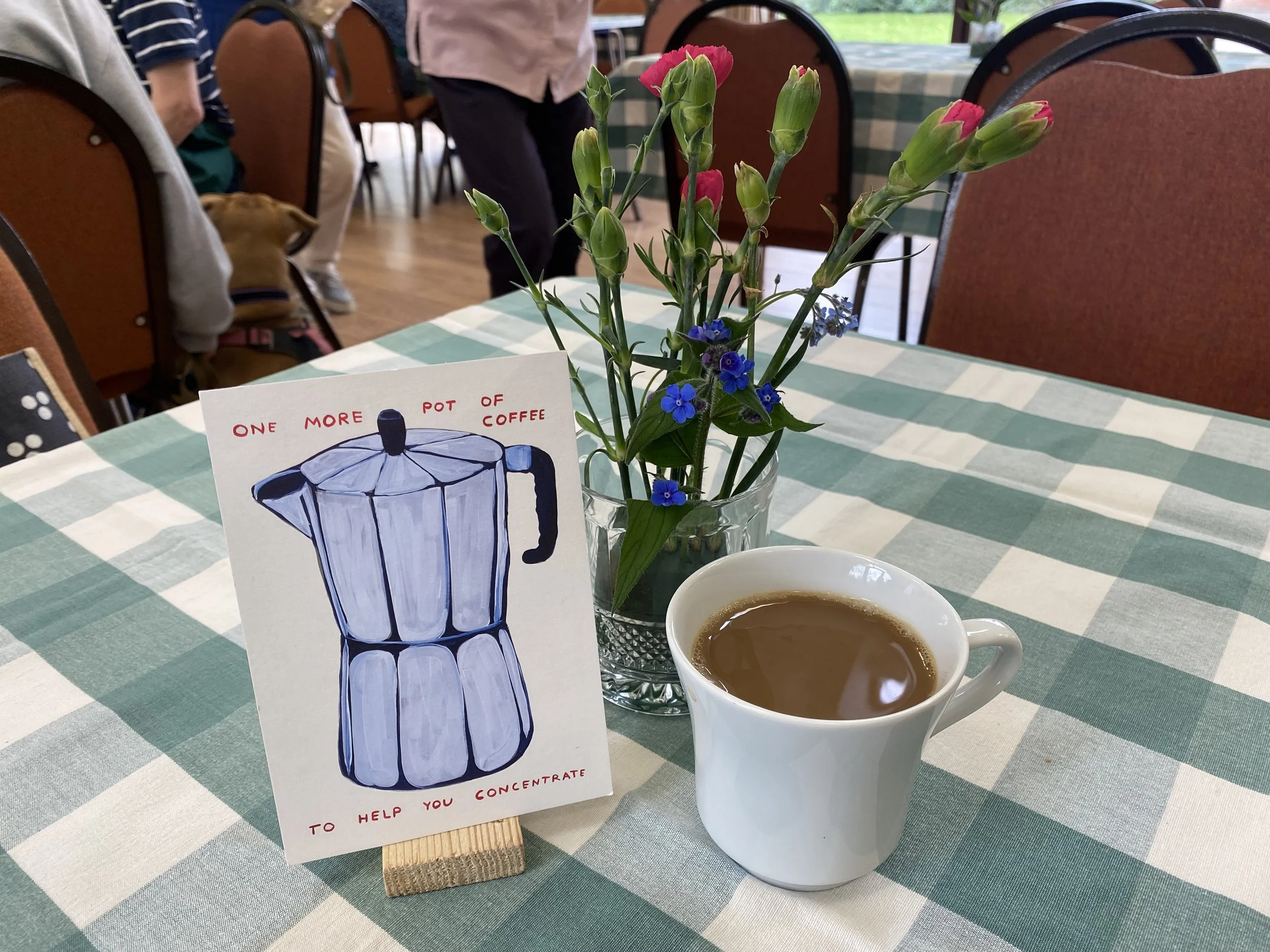 A table with a cup of coffee, a small vase of pink and purple flowers, and a card featuring a drawing of a moka pot with the caption "One more pot of coffee to help you concentrate." The table is covered with a green and white checkered tablecloth, a