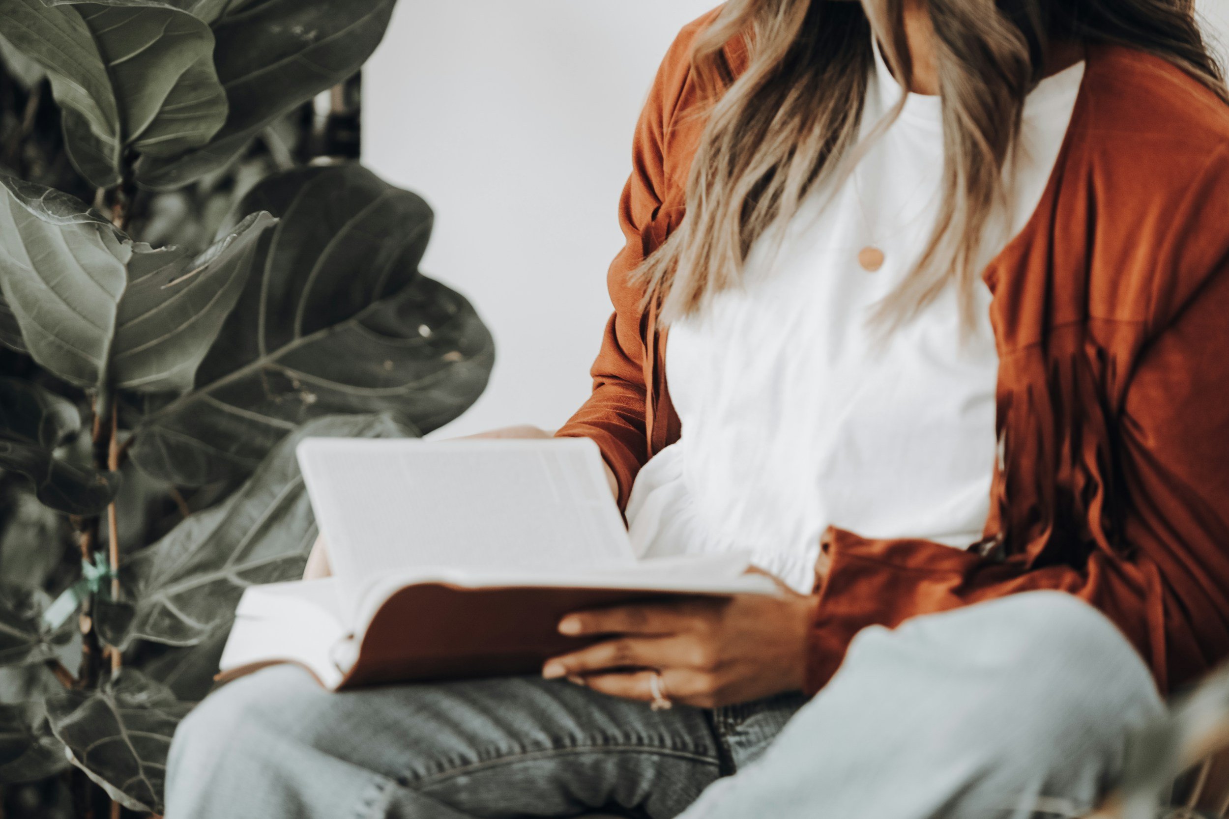 A person sitting and reading a book next to a large indoor plant, wearing a white shirt and a brown jacket.