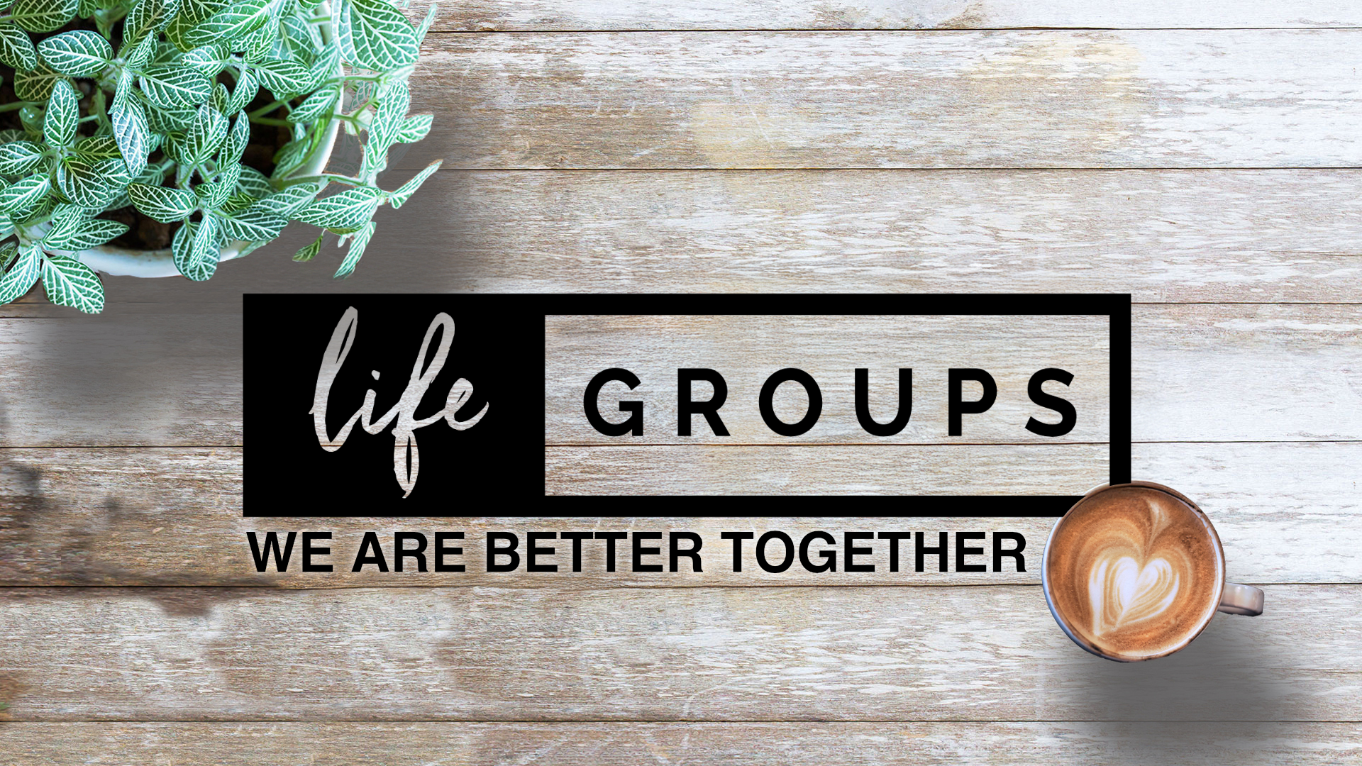 A sign that reads 'life groups, we are better together' with a potted plant on the top left corner and a coffee mug with a heart latte art on the bottom right, placed on a wooden table.