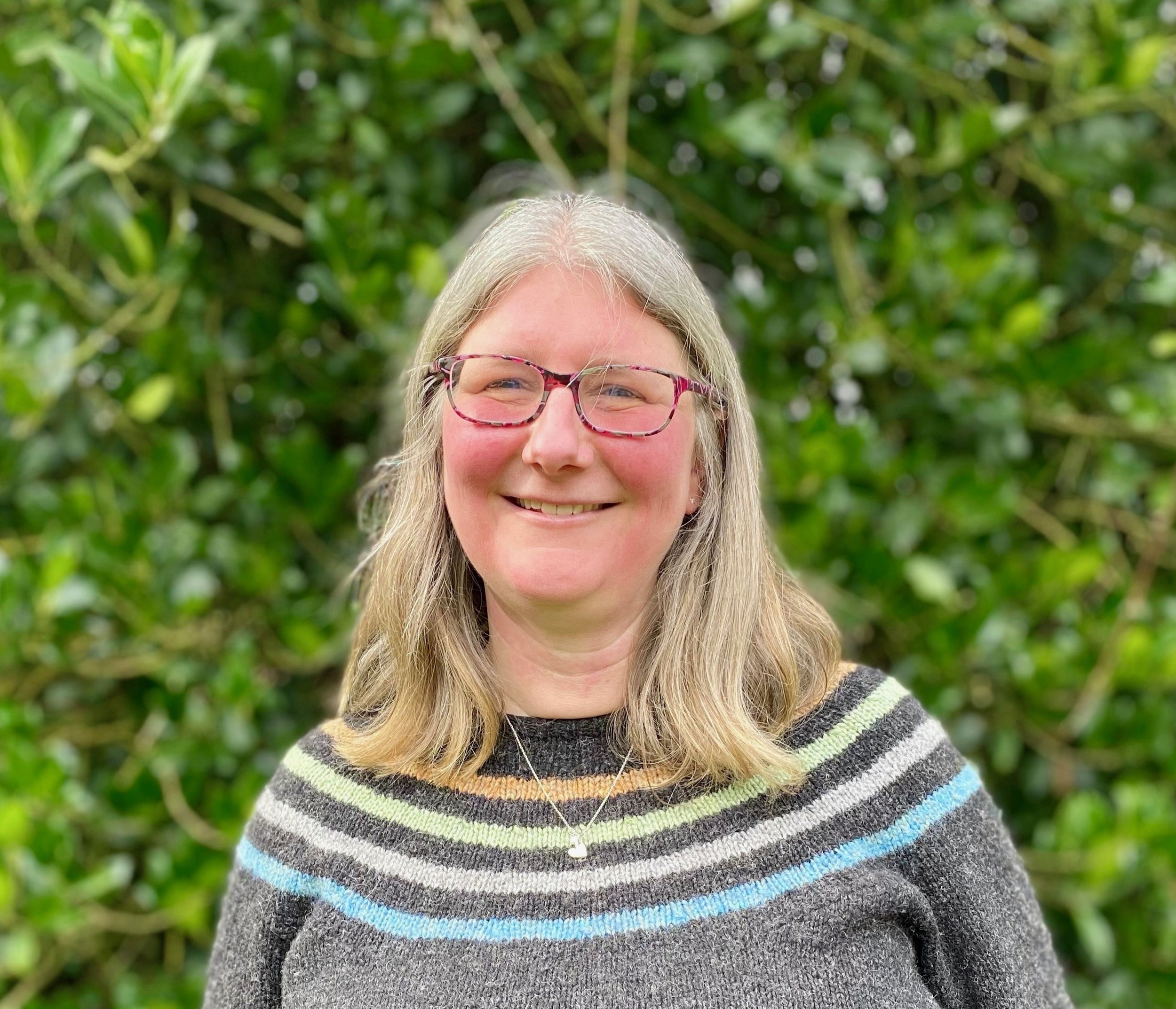 A woman with shoulder-length gray hair, wearing glasses and a multicolored striped sweater, smiling outdoors with green foliage in the background.