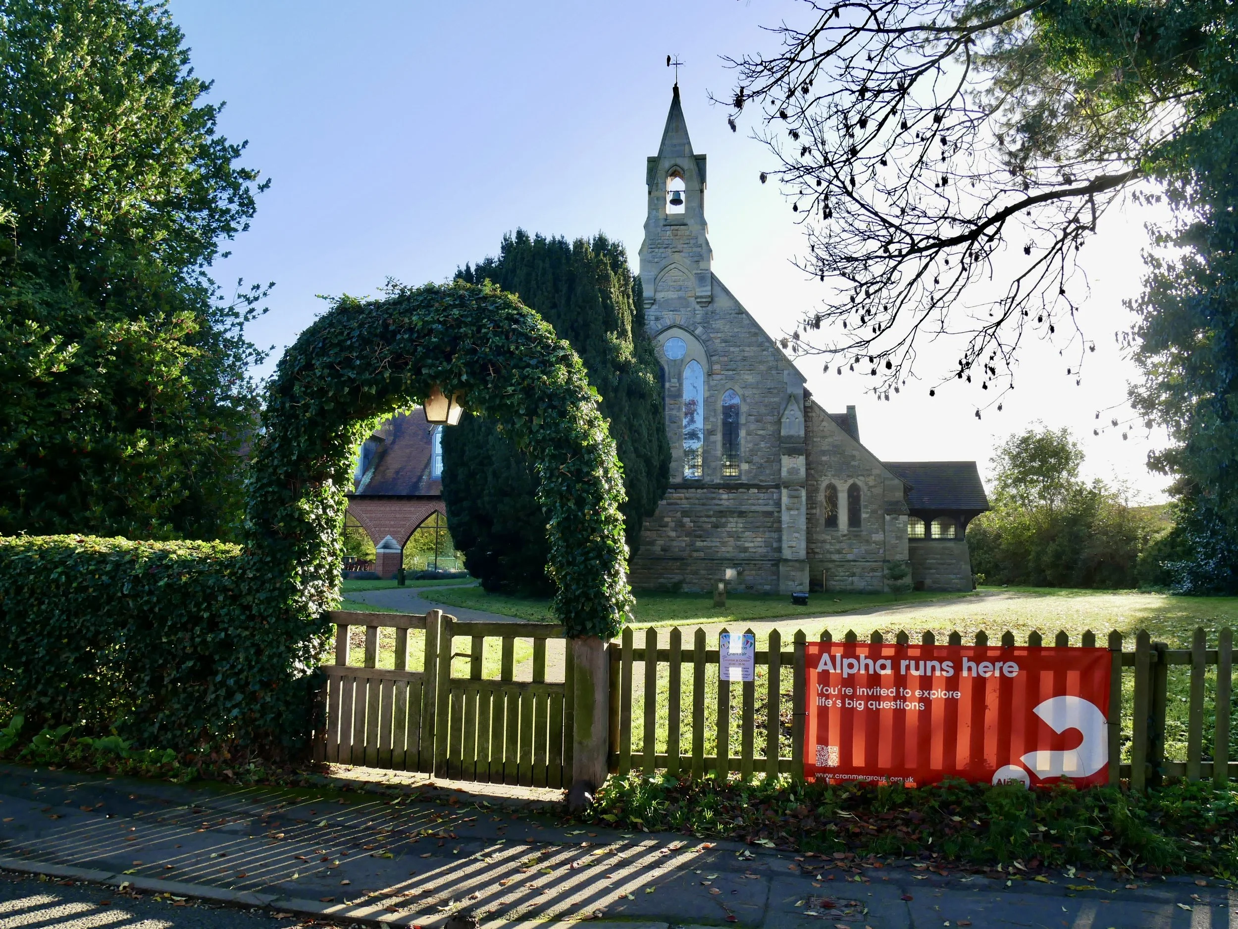 A church with a stone facade viewed through a wooden fence in a park, with trees and a clear blue sky in the background. A red banner reads 'Alpha runs here' inviting exploration of life's big questions.