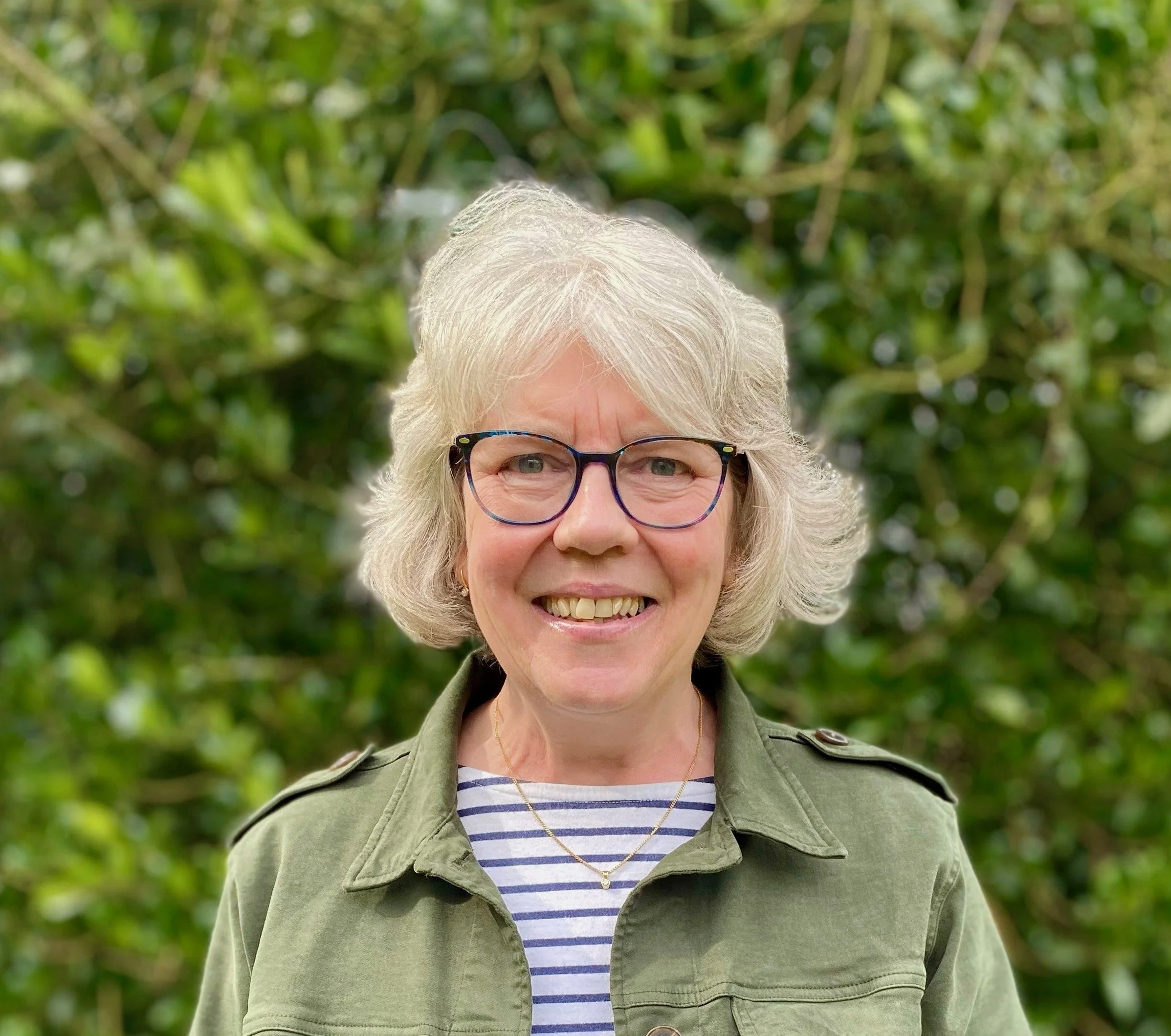 Smiling elderly woman with glasses, short gray hair, wearing a green jacket over a striped shirt, standing outdoors with green foliage in the background.