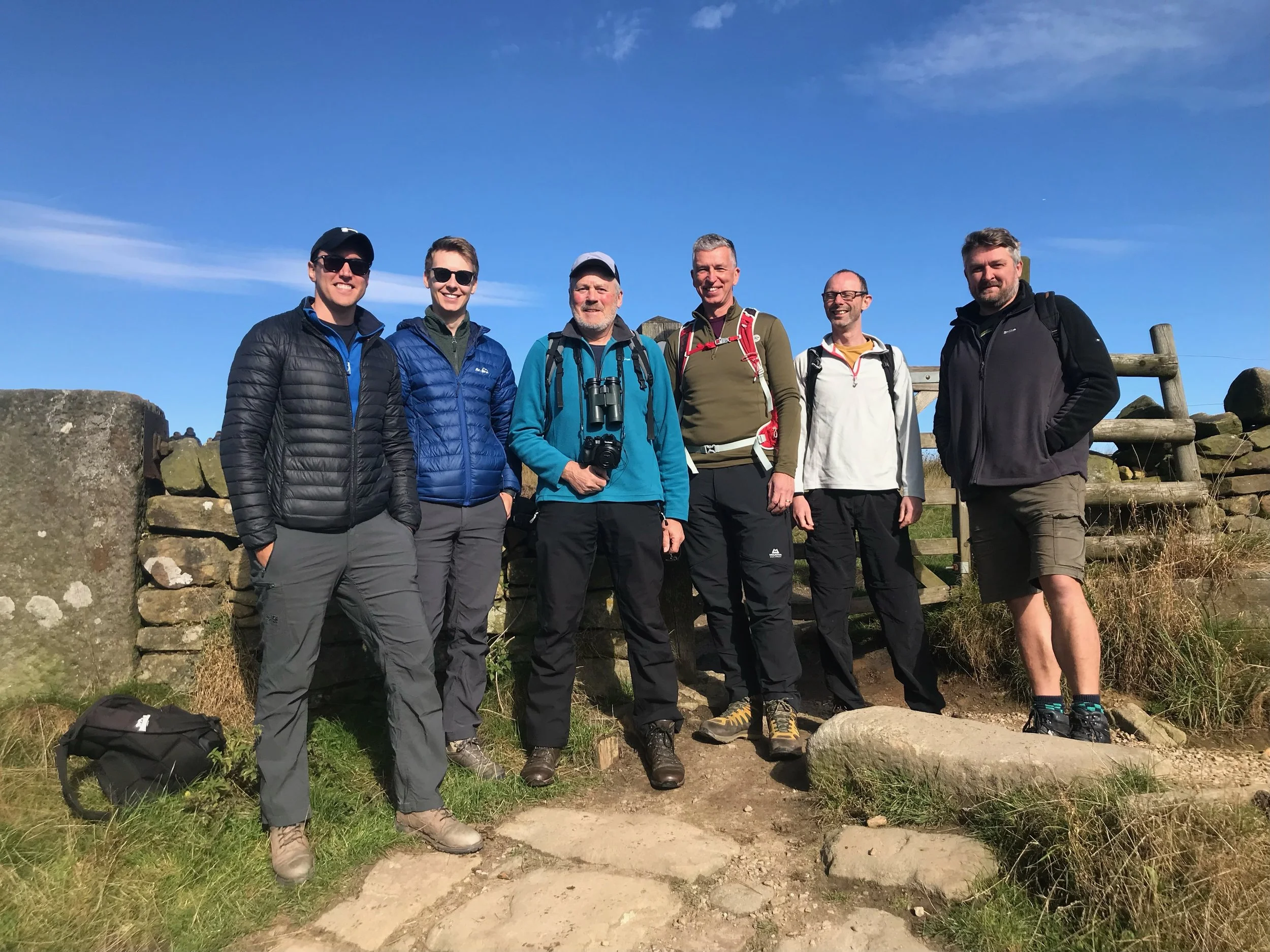 Six men hiking outdoors on a sunny day, standing in front of a stone fence with grass and rocks around, wearing outdoor gear and backpacks.