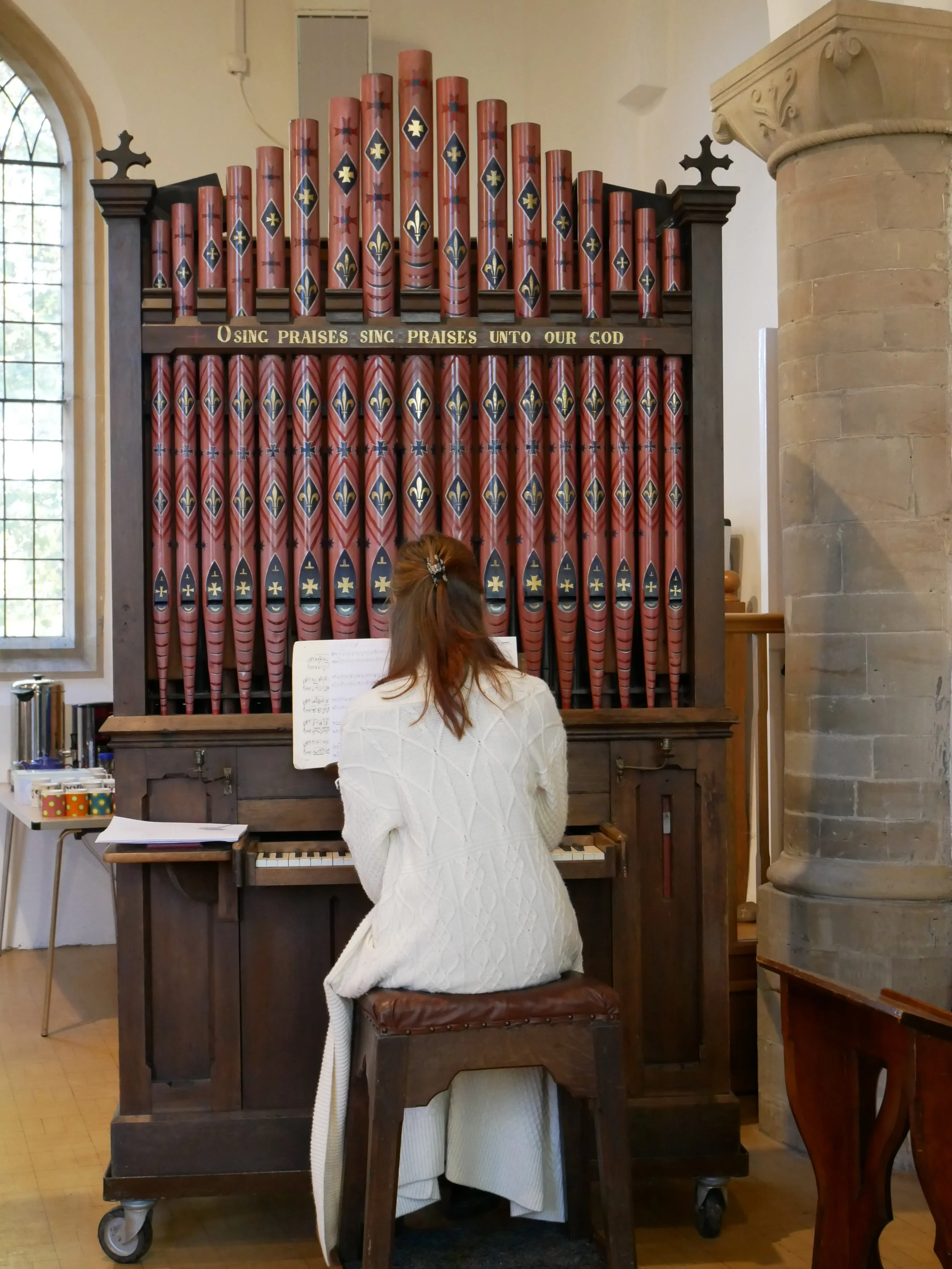 A person with long red hair in a white sweater playing an organ in a church, with the words 'O SING PRAISES SING PRAISES UNTO OUR GOD' written on the organ.