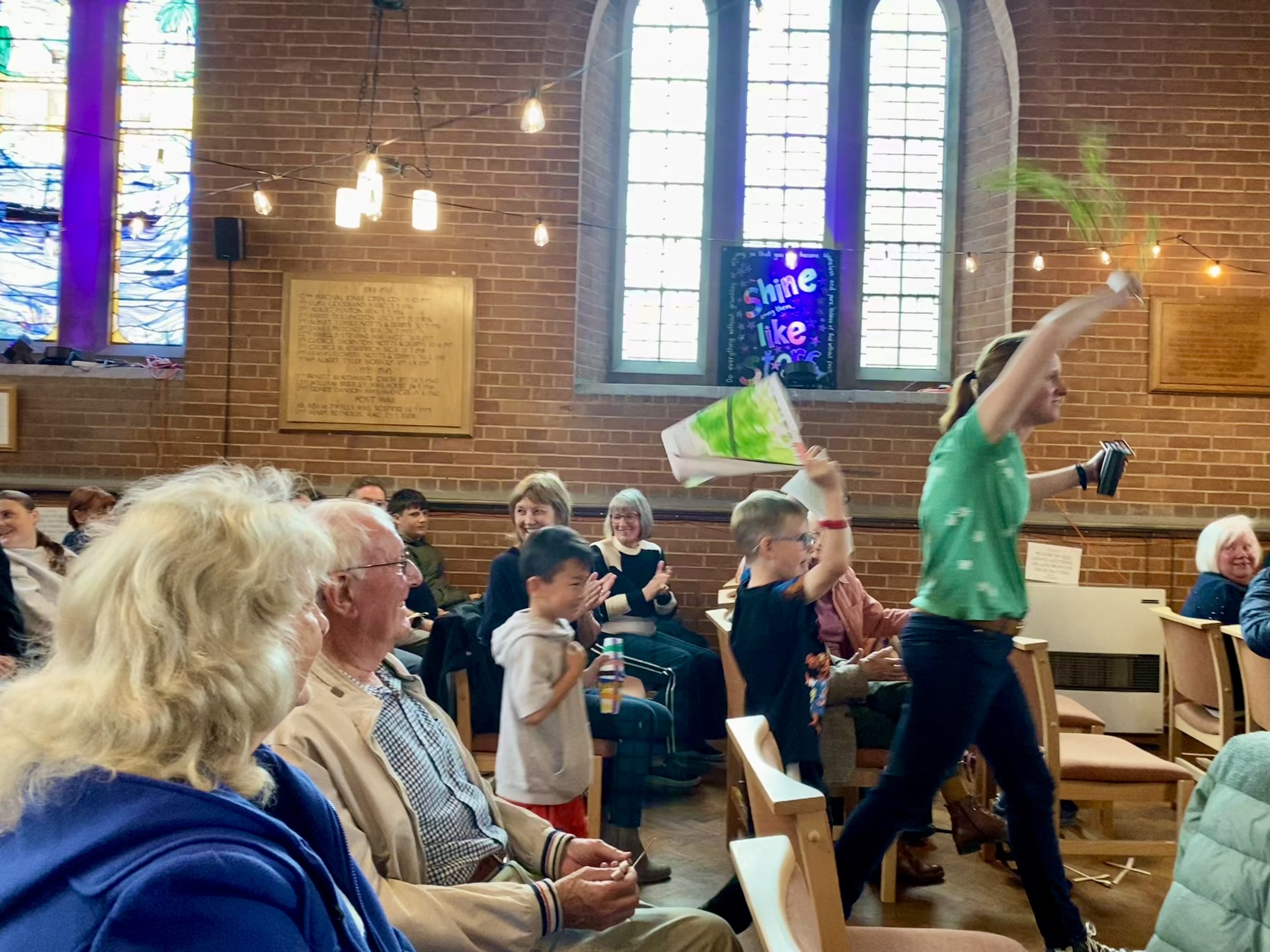 A woman in a green shirt is walking through a church with an audience of children and adults seated on chairs. The woman is waving a colorful, cone-shaped flag and holding a notebook. The background shows stained glass windows and a brick wall with plaques.