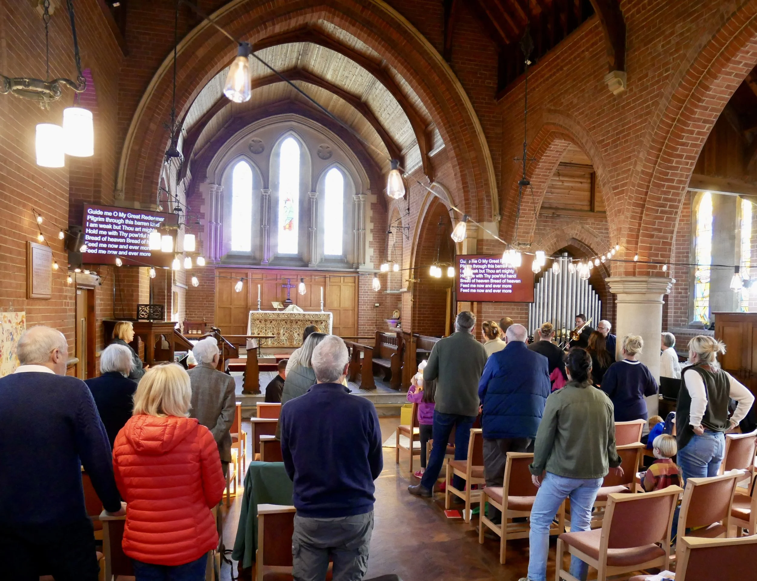 People gathered inside a church for a service or event, with stained glass windows, an altar, and a pipe organ in the background, illuminated by hanging lights.