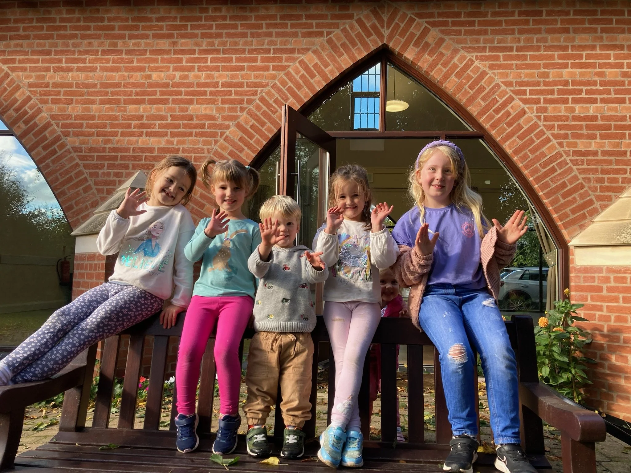 Six children, five girls and one boy, sit on a wooden bench outside a brick building with large, arched windows. They are smiling and waving at the camera.