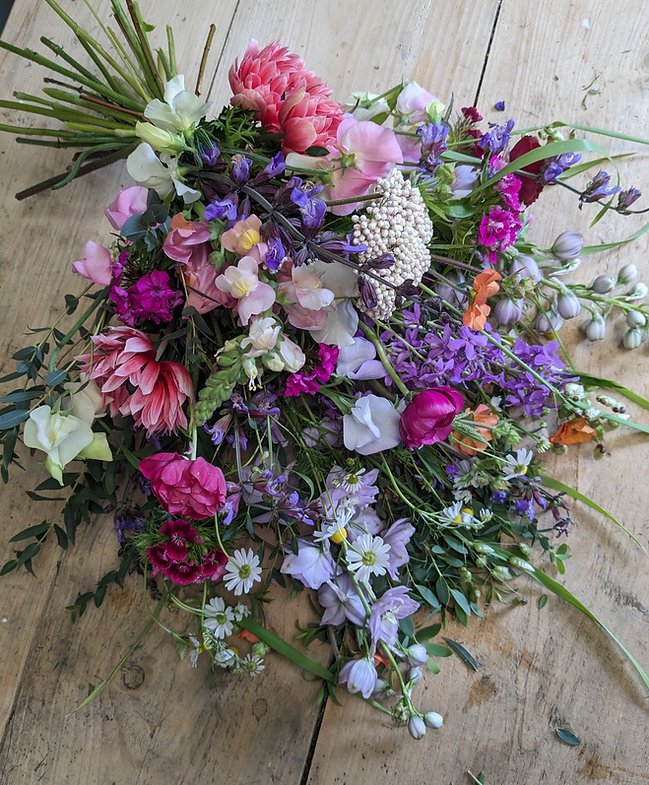 A colorful mixed bouquet of flowers including pink, purple, white, and orange blooms, arranged with greenery, resting on a wooden surface.