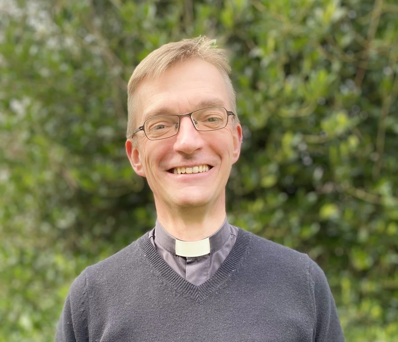 A smiling man with glasses, wearing a clerical collar and dark sweater, standing outdoors with trees in the background.
