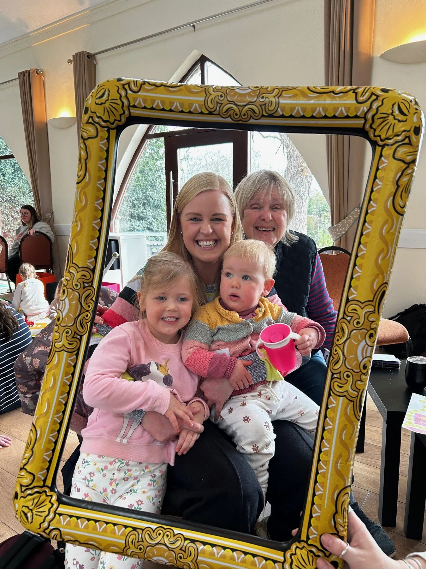 Group of four family members smiling and sitting together, framed within a decorative yellow picture frame, in a room with large windows and curtains.
