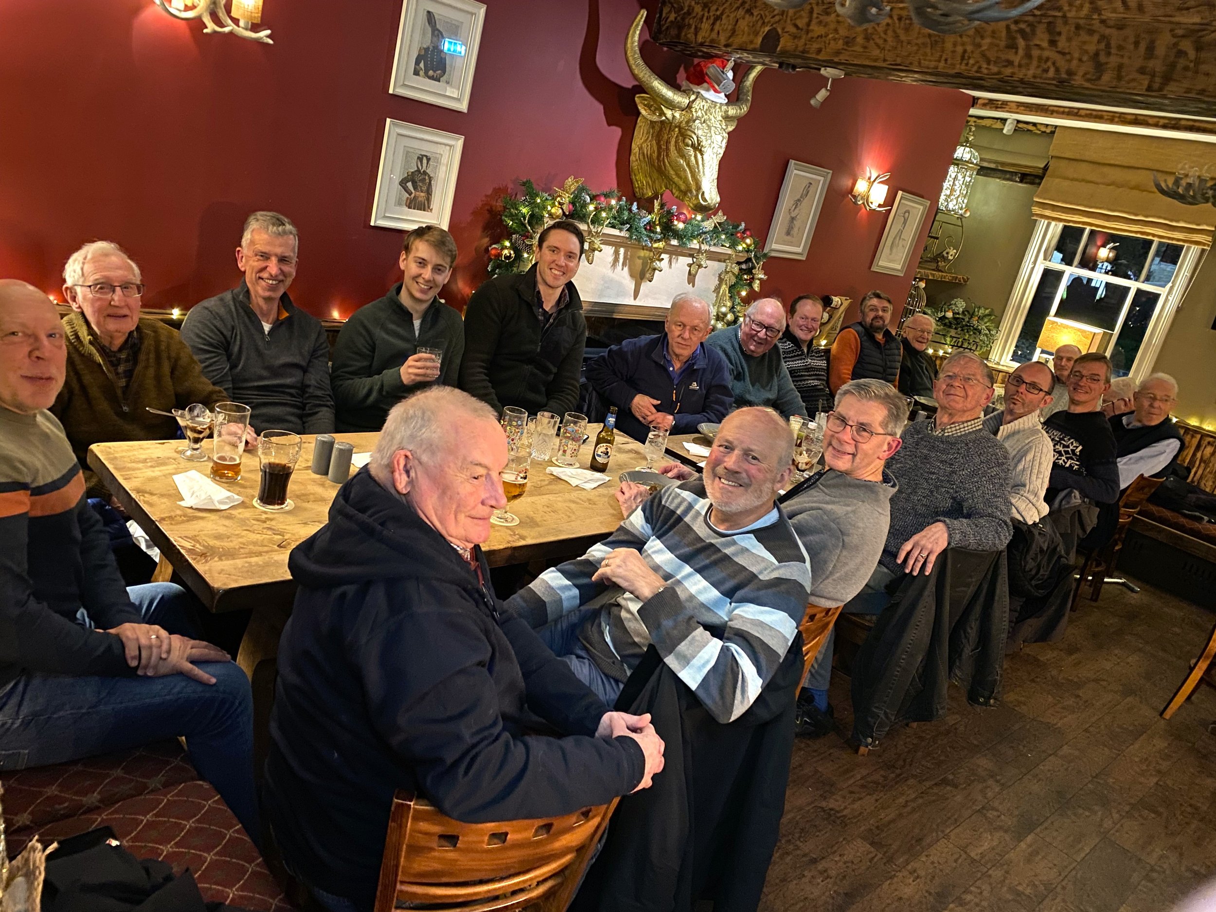 A group of 16 men gathered around a large wooden table in a warmly lit room decorated with Christmas ornaments, a Christmas tree, and a mounted animal head above the fireplace.