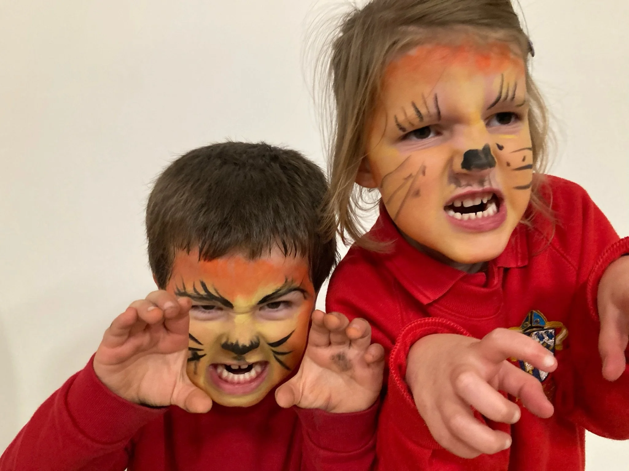 Two children with painted faces dressed as animals, one with a tiger face and the other with a lion face, making playful clawing gestures.