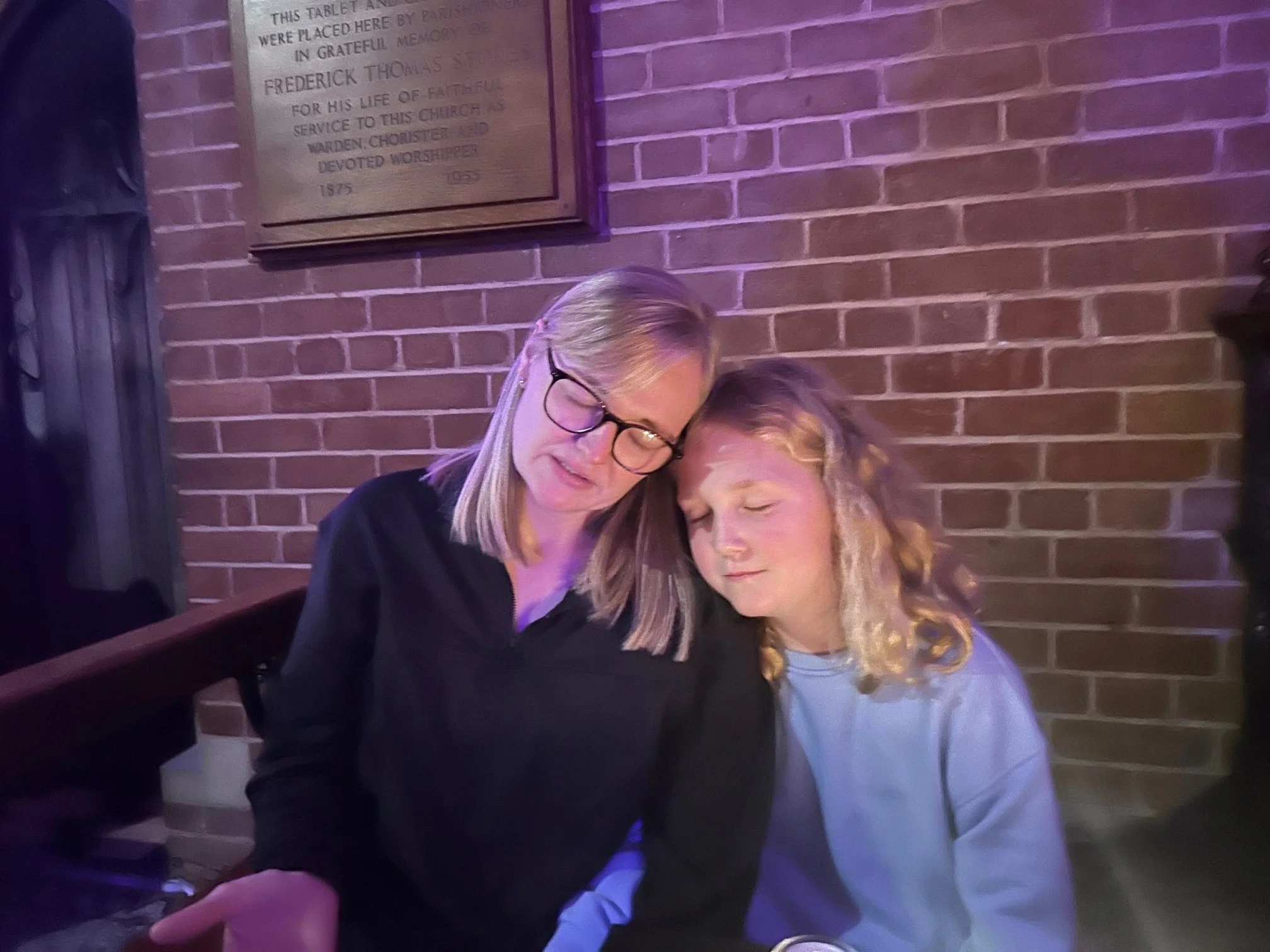 A woman with glasses and a girl with curly blonde hair sit close together with their heads resting against each other in a dimly lit room with a brick wall backdrop.