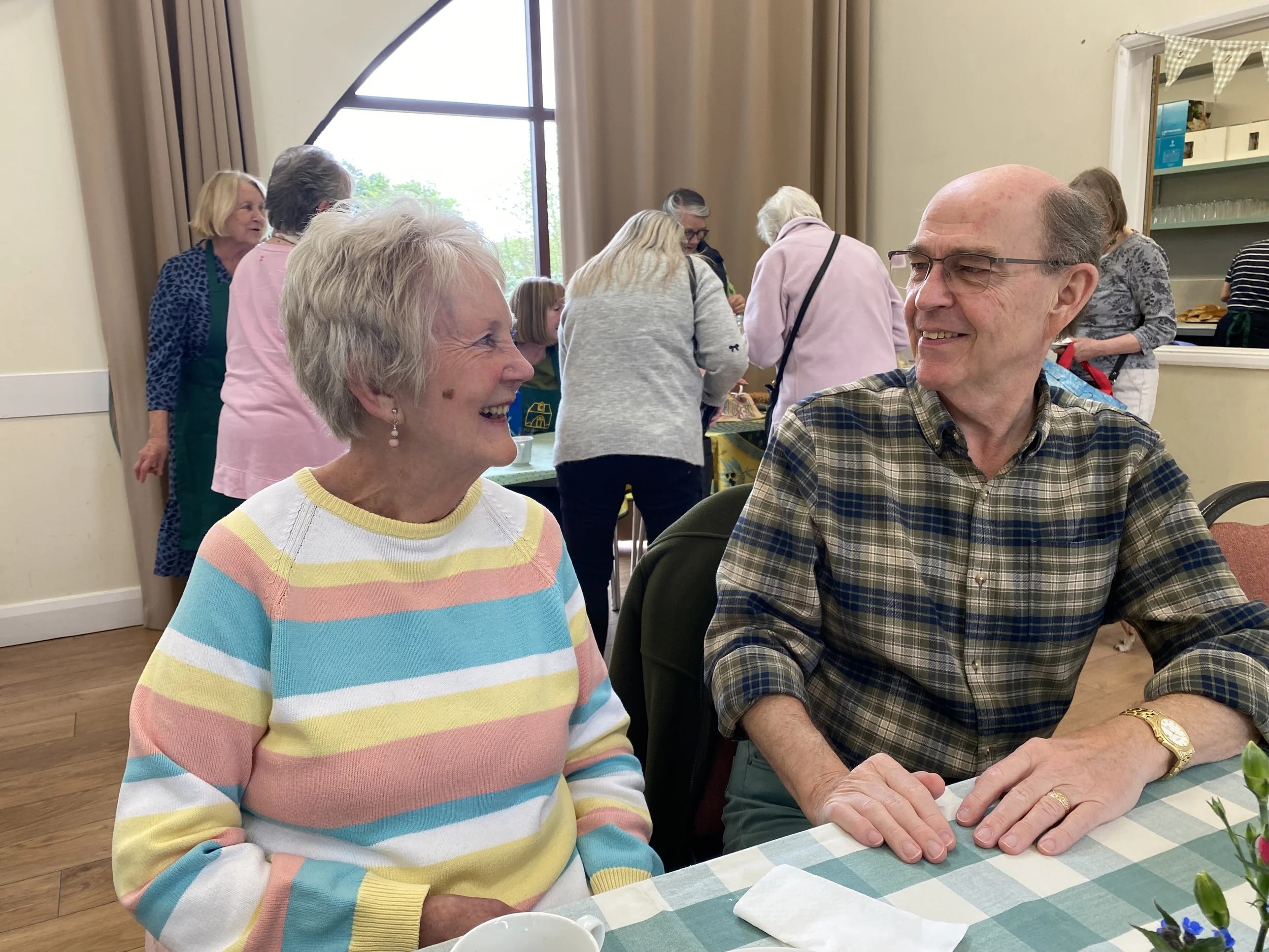 An elderly woman and a man smiling and enjoying conversation at a gathering in a room with other people in the background.