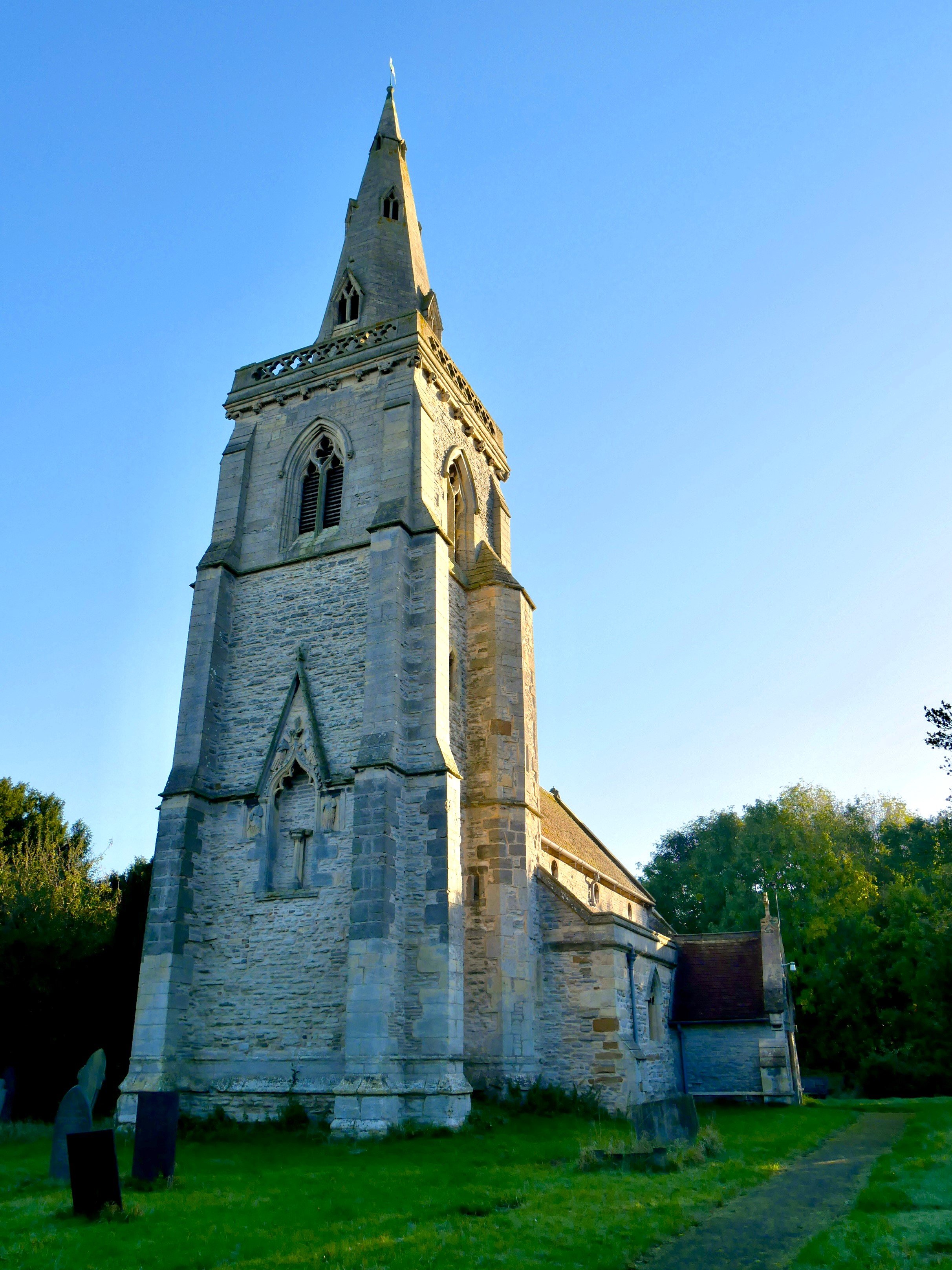 A stone church with a tall steeple against a clear blue sky. The church is surrounded by green grass and trees.