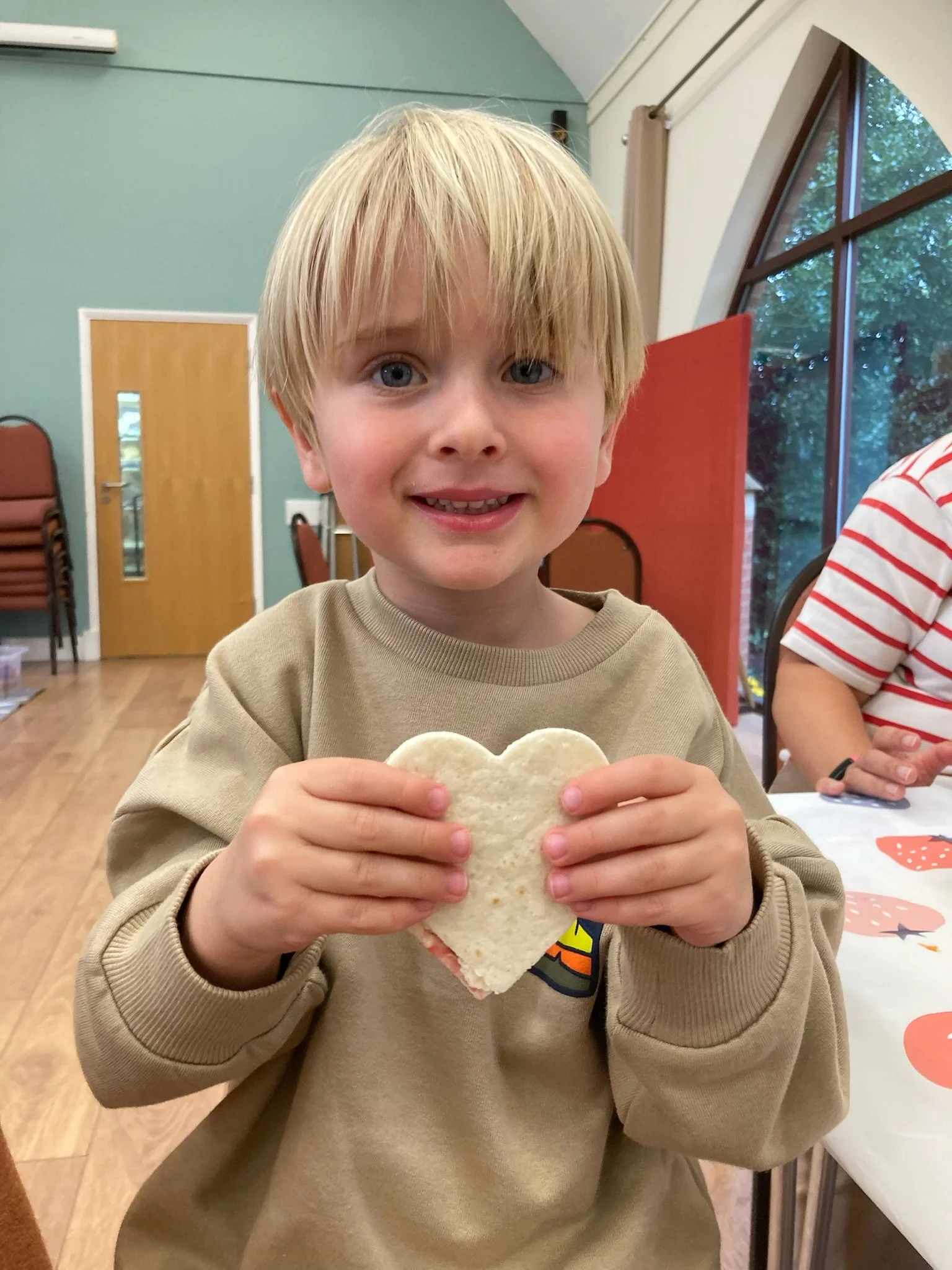 A young boy  smiling while holding a heart-shaped piece of bread or cookie at a table in a bright room.