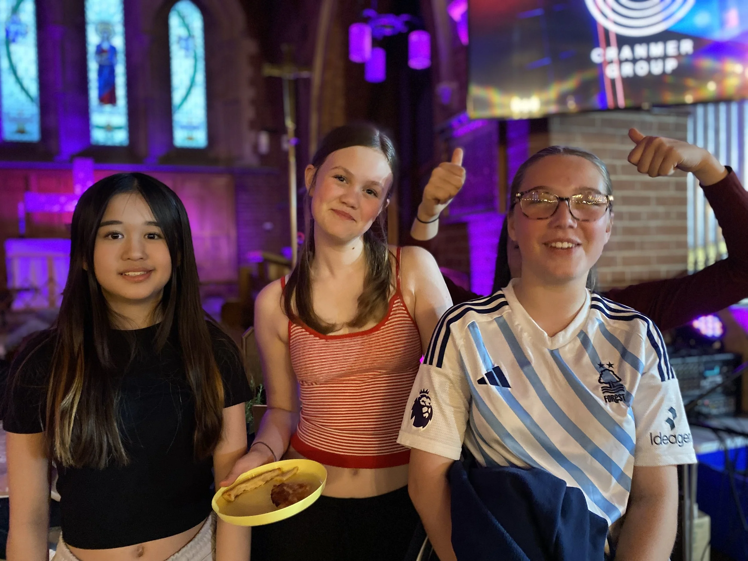 Three young women stand indoors, with a brick wall and stained glass windows behind them, under colorful lighting. The woman on the right, wearing glasses and a soccer jersey, is smiling and making a fist pump gesture. The woman in the middle, wearing a tank top, is holding a yellow plate with food and smiling. The woman on the left, in a black crop top, is smiling as well.