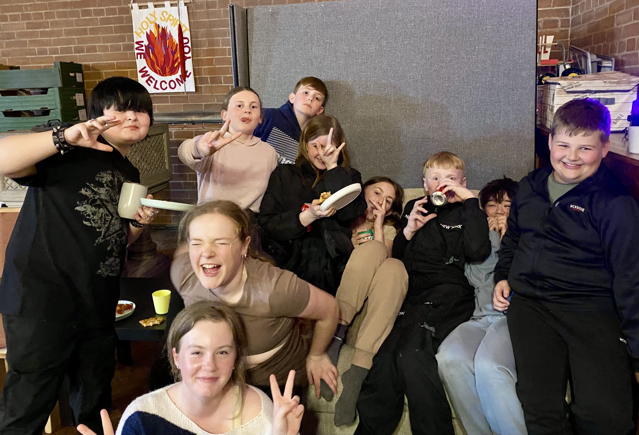 Group of kids enjoying a casual gathering indoors, some making peace signs, eating, and laughing, with a brick wall and poster in background.