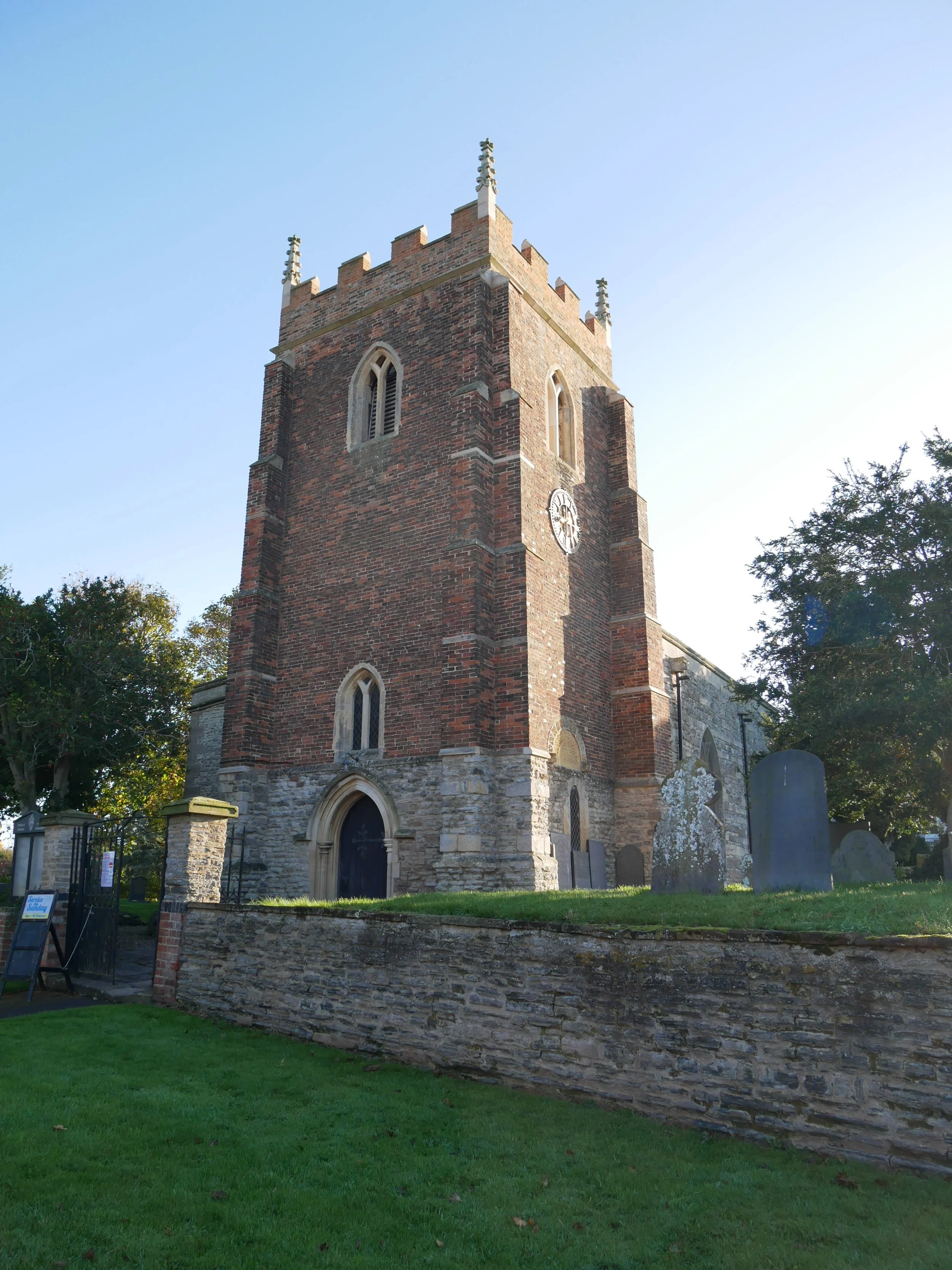A tall brick church tower with gothic windows and a clock, surrounded by a stone wall and graveyard, under a clear blue sky.
