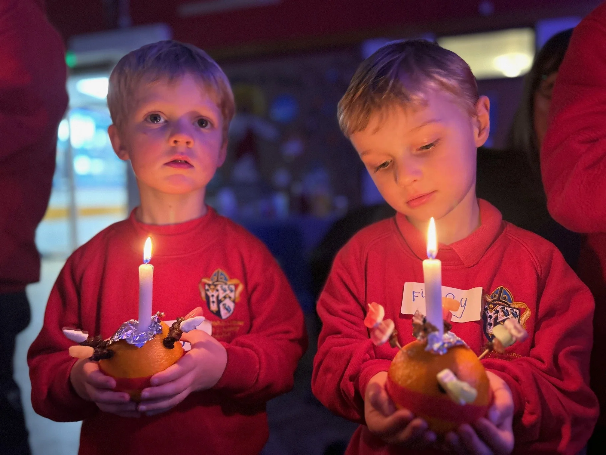 Two young boys wearing red shirts with a crest, holding oranges with lit birthday candles and decorated with foil and small objects, standing in a dimly lit environment, possibly celebrating a birthday.