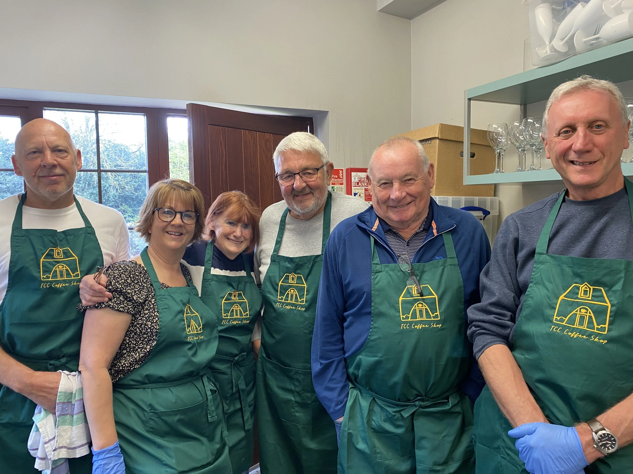 A group of six smiling people standing inside a kitchen, wearing green aprons with a yellow logo that says 'TCC Coffee Shop.' They are posing together, some with glasses or gloves, in front of wooden and metal shelves with glassware.