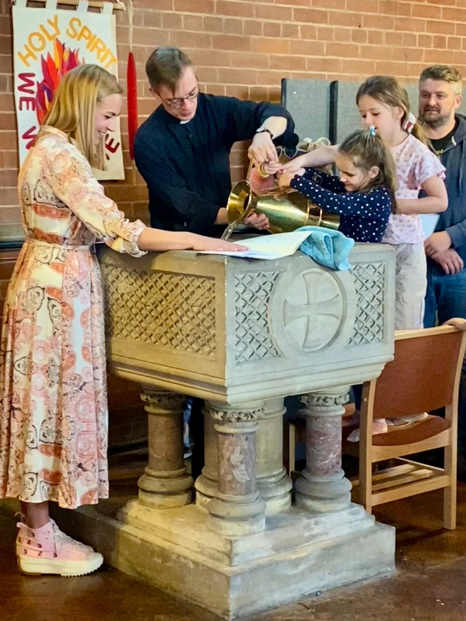 A group of people, including a woman, a priest, and children, are gathered around a baptismal font in a church. The priest is pouring water over a child's arm during a baptism ceremony. The setting includes a brick wall, a banner that says 'Holy Spirit We Welcome You,' and church furnishings.