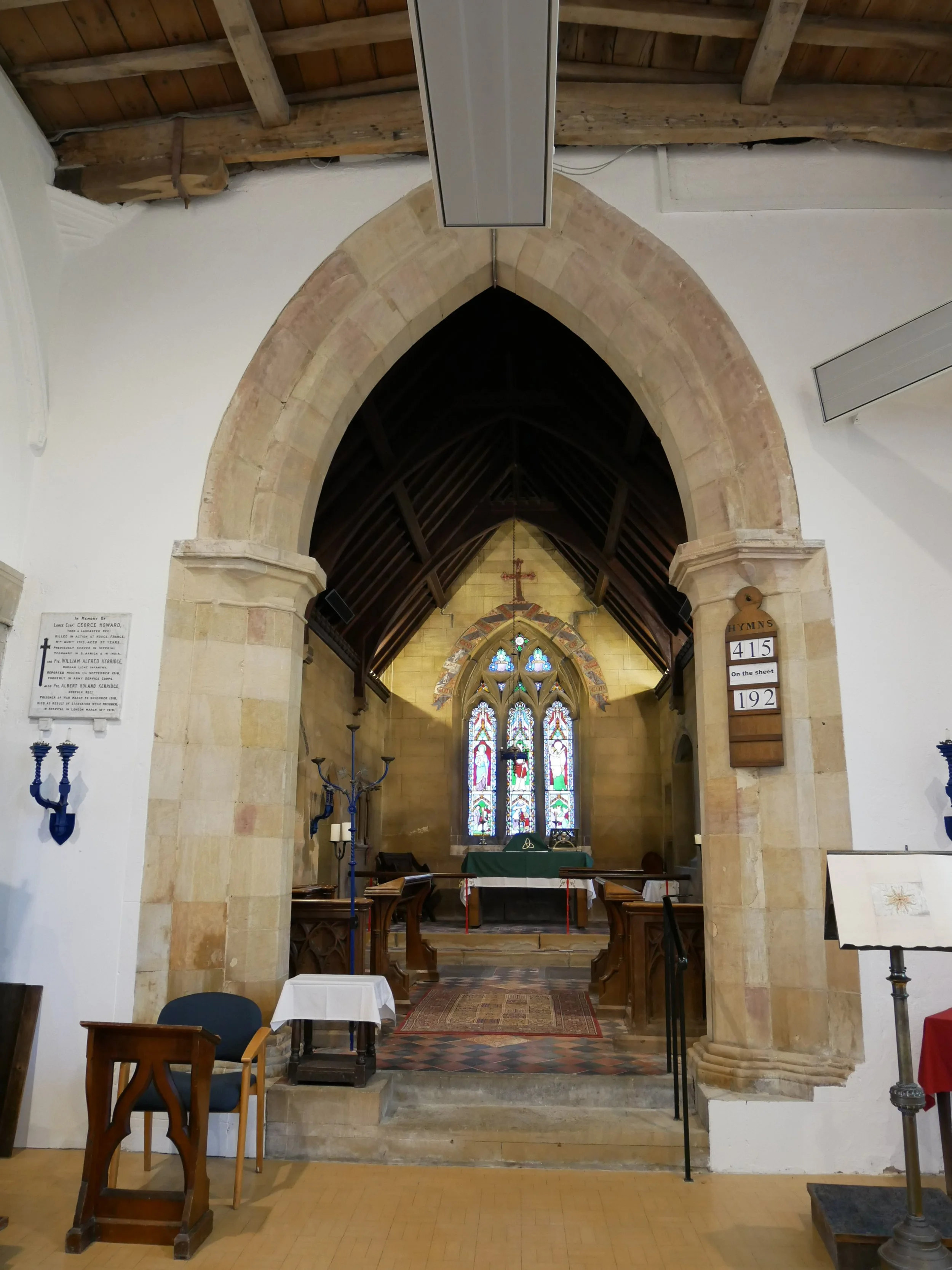 Interior of a church with an arched stone doorway leading to the altar area, which has stained glass windows and an altar table with liturgical items. There are pews, chairs, and candelabras inside.
