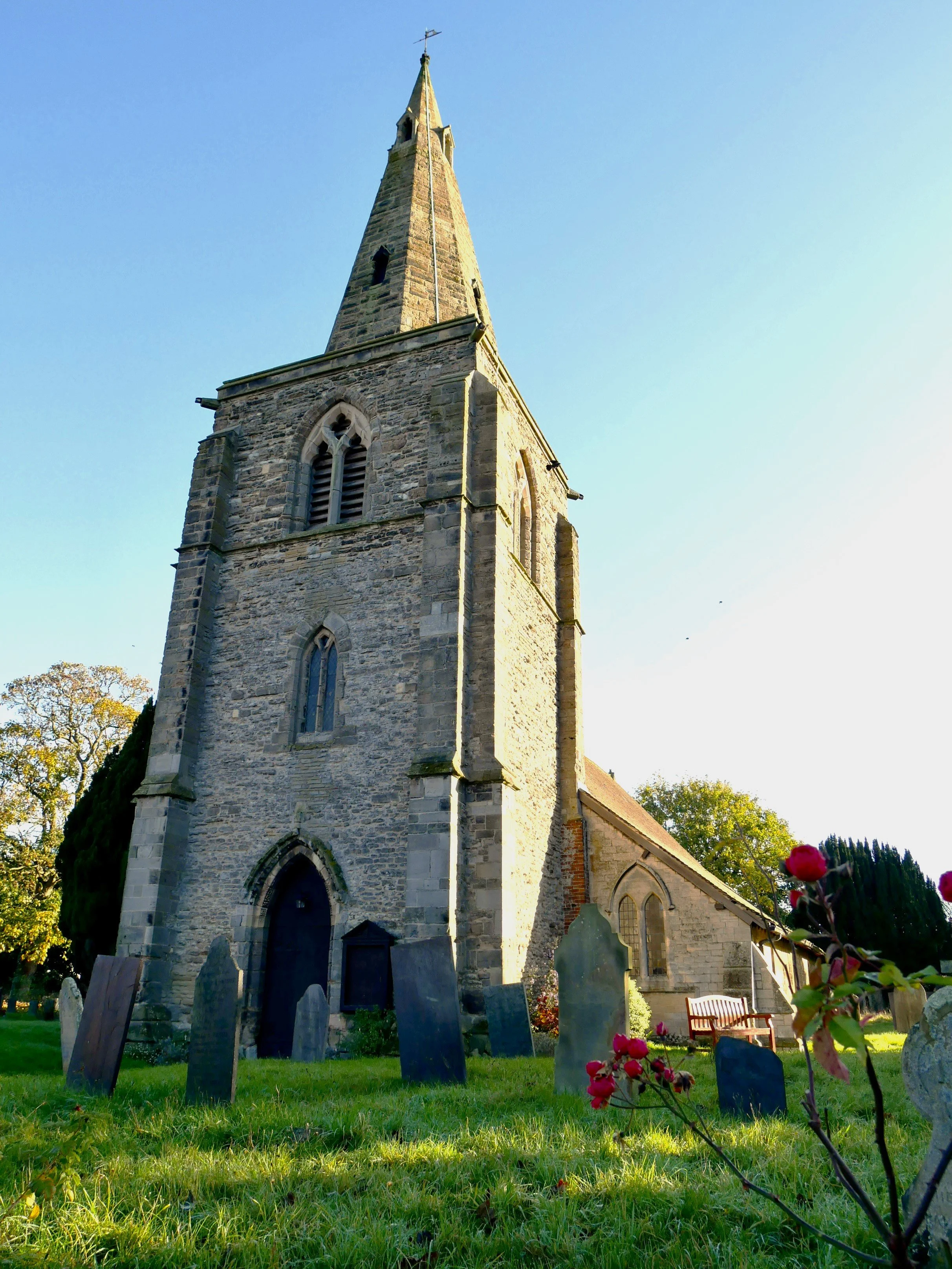 A tall stone church with a pointed steeple and tall narrow windows, surrounded by a graveyard with headstones and a bench under trees, during daytime with clear sky.