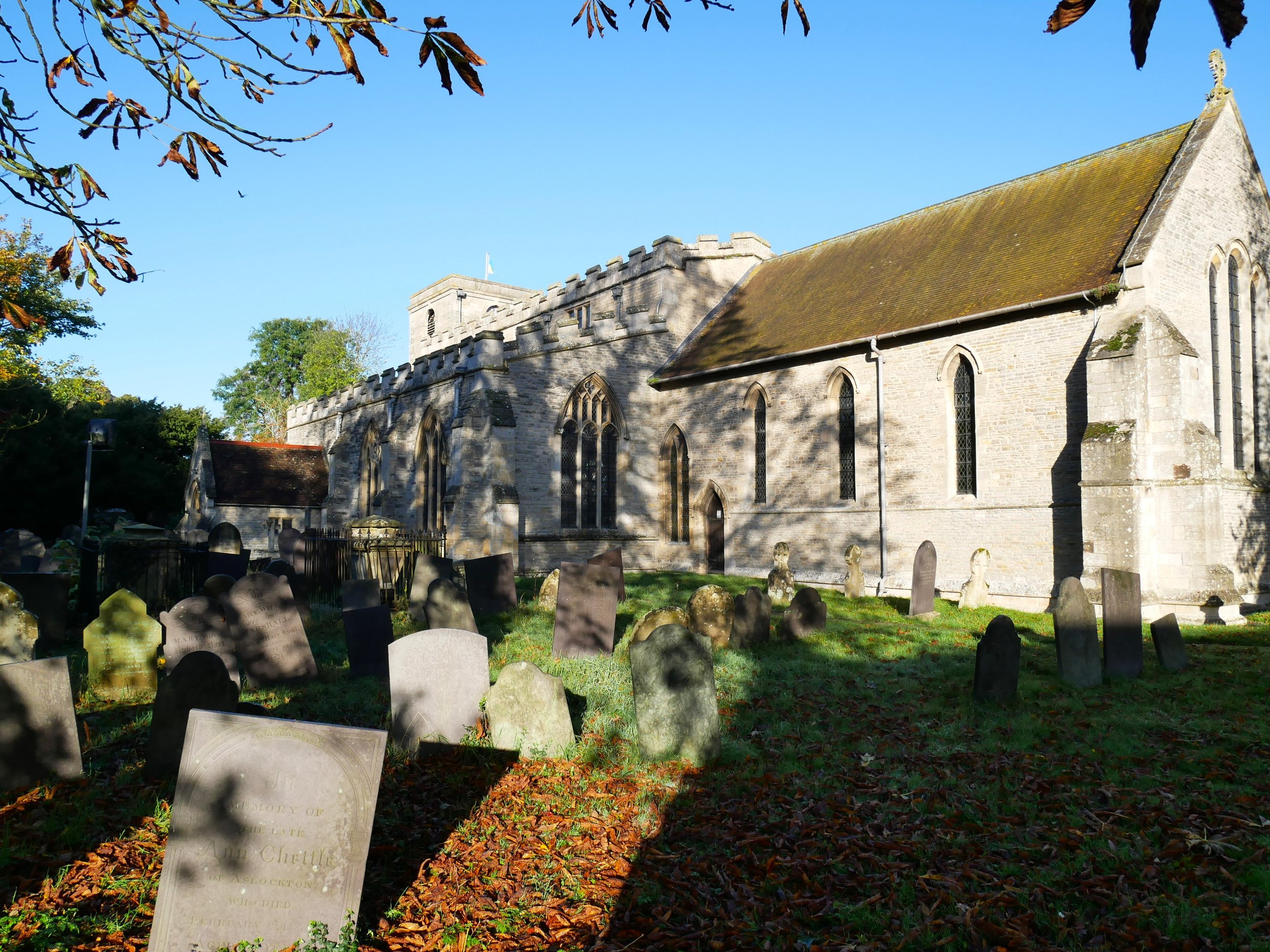 A historic stone church with tall arched windows and a graveyard filled with headstones in the foreground, some of which are shaded by tree branches. The sky is clear and sunny, casting shadows on the church and the cemetery.