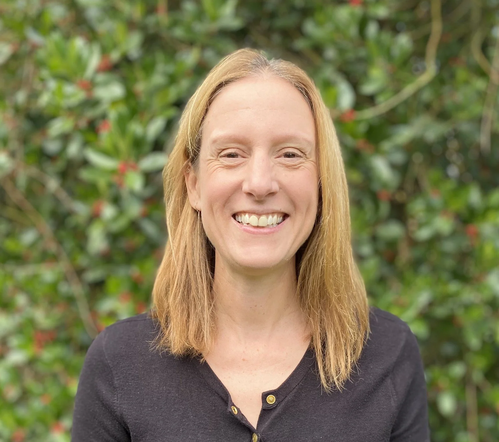 A woman with shoulder-length blonde hair smiling outdoors with greenery background.