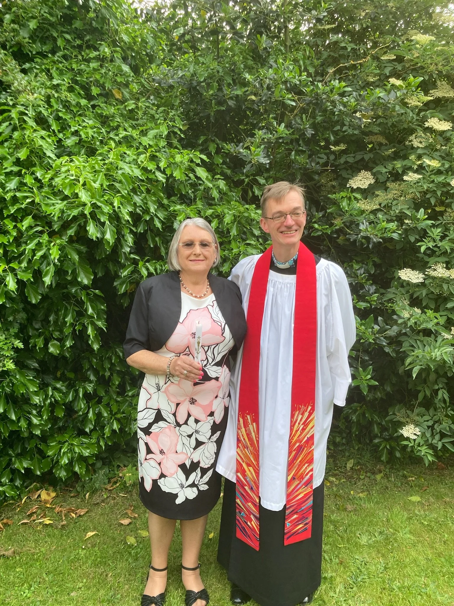 A woman and a man standing outdoors in front of green bushes. The woman is holding a candle, and the man is wearing a white clergy robe with a red stole. They are smiling and appear to be at a religious or ceremonial event.