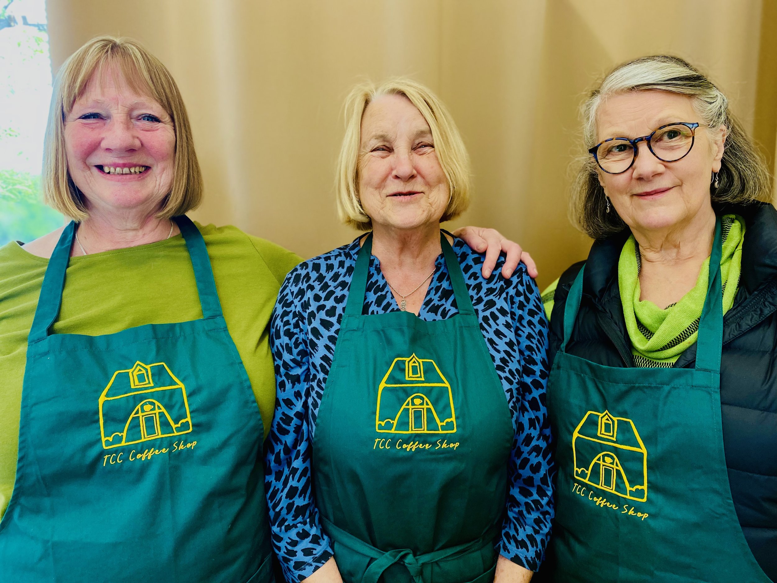 Three older women wearing teal aprons with a yellow house logo and the text 'TCC Coffee Shop' pose together indoors. They smile, and the woman in the middle has her arm around the woman on the right. The background includes beige curtains.