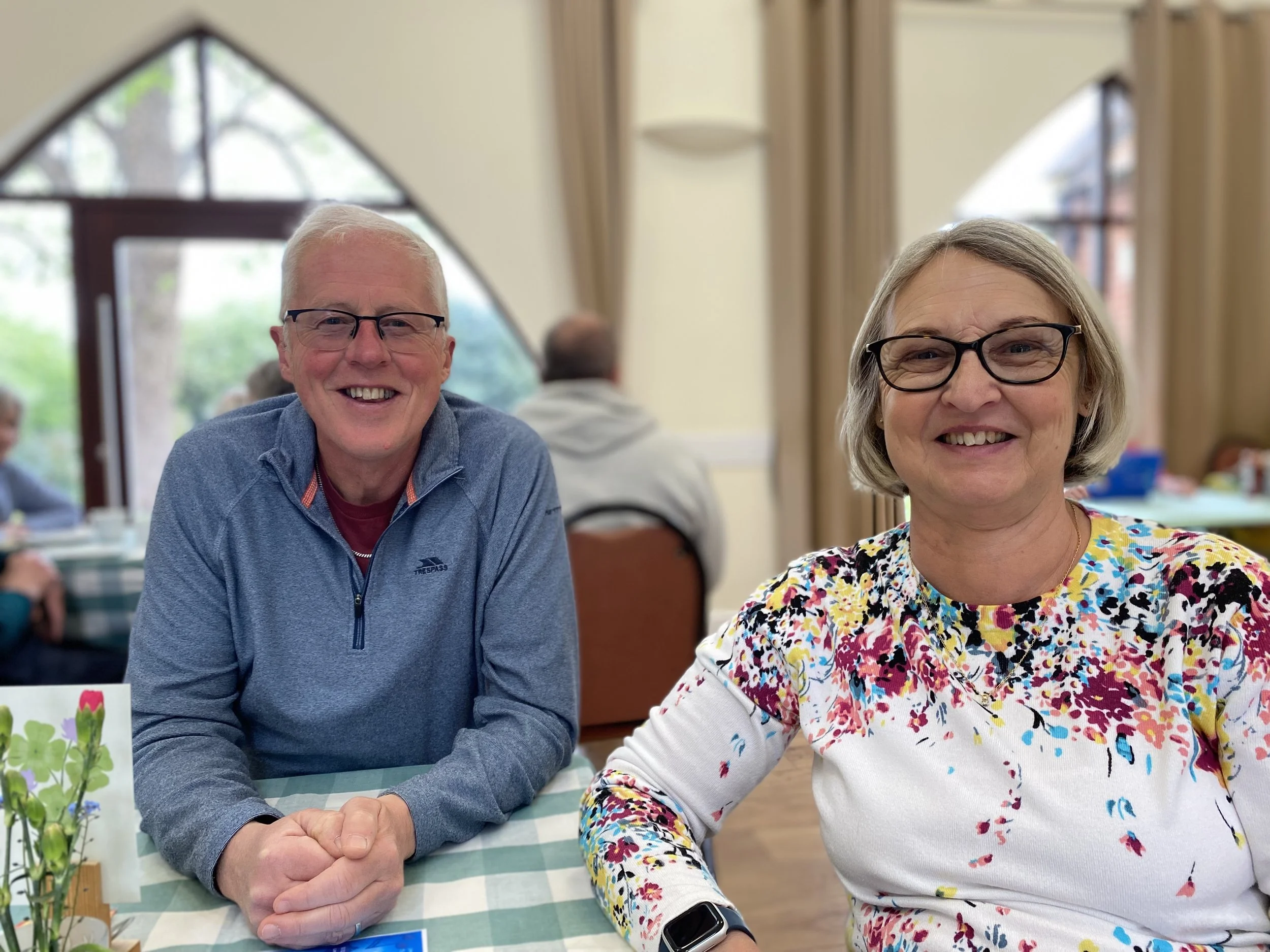 An older man and woman smiling at a table in a bright room with large arched windows. The man has short white hair, glasses, and is wearing a blue zip-up jacket. The woman has short gray hair, glasses, and is wearing a colorful floral top. There are 
