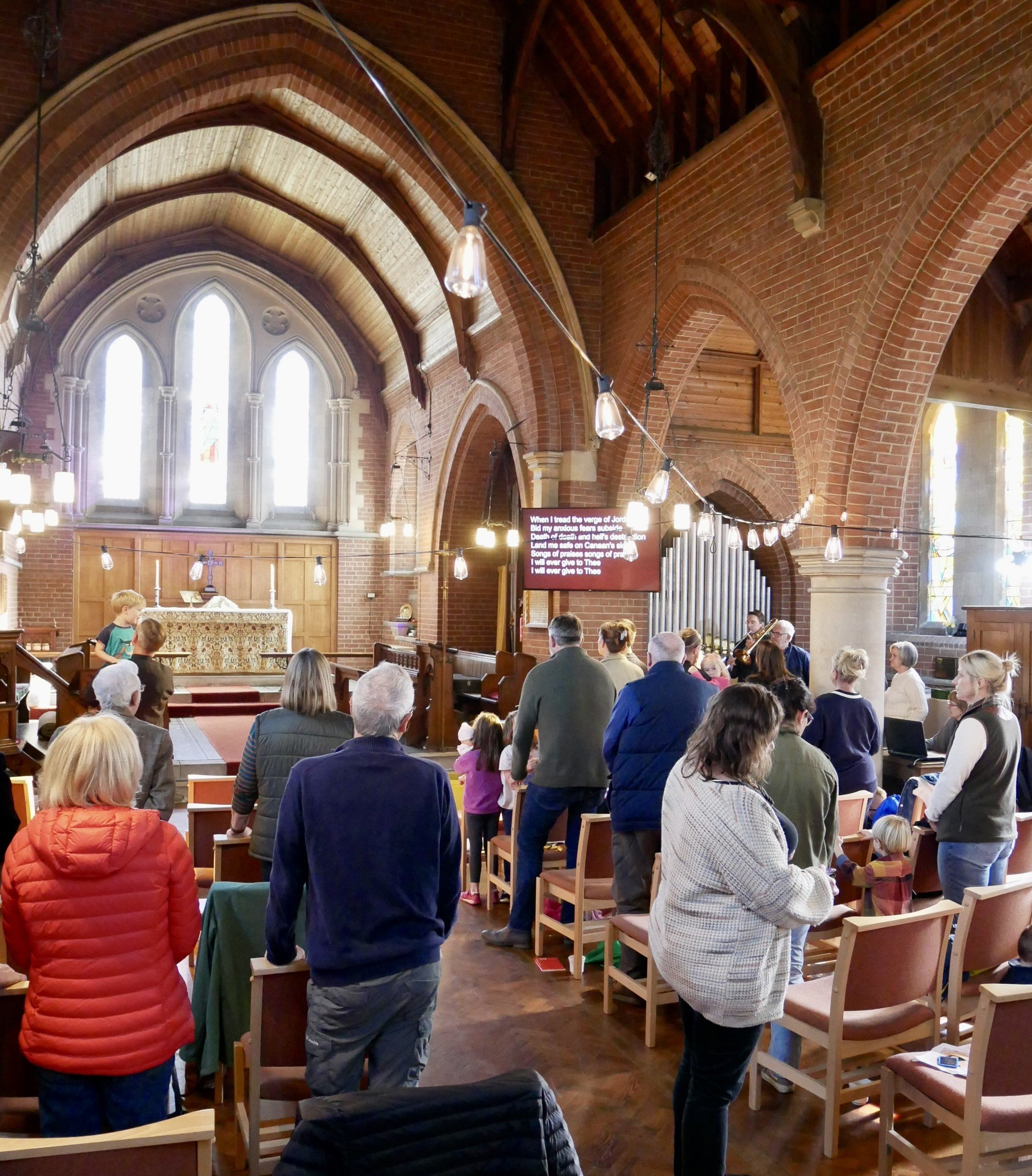 People gathered inside a church during a service or event, with some standing and some seated, facing the altar at the front. The church has stained glass windows, brick walls, and wooden architectural features.