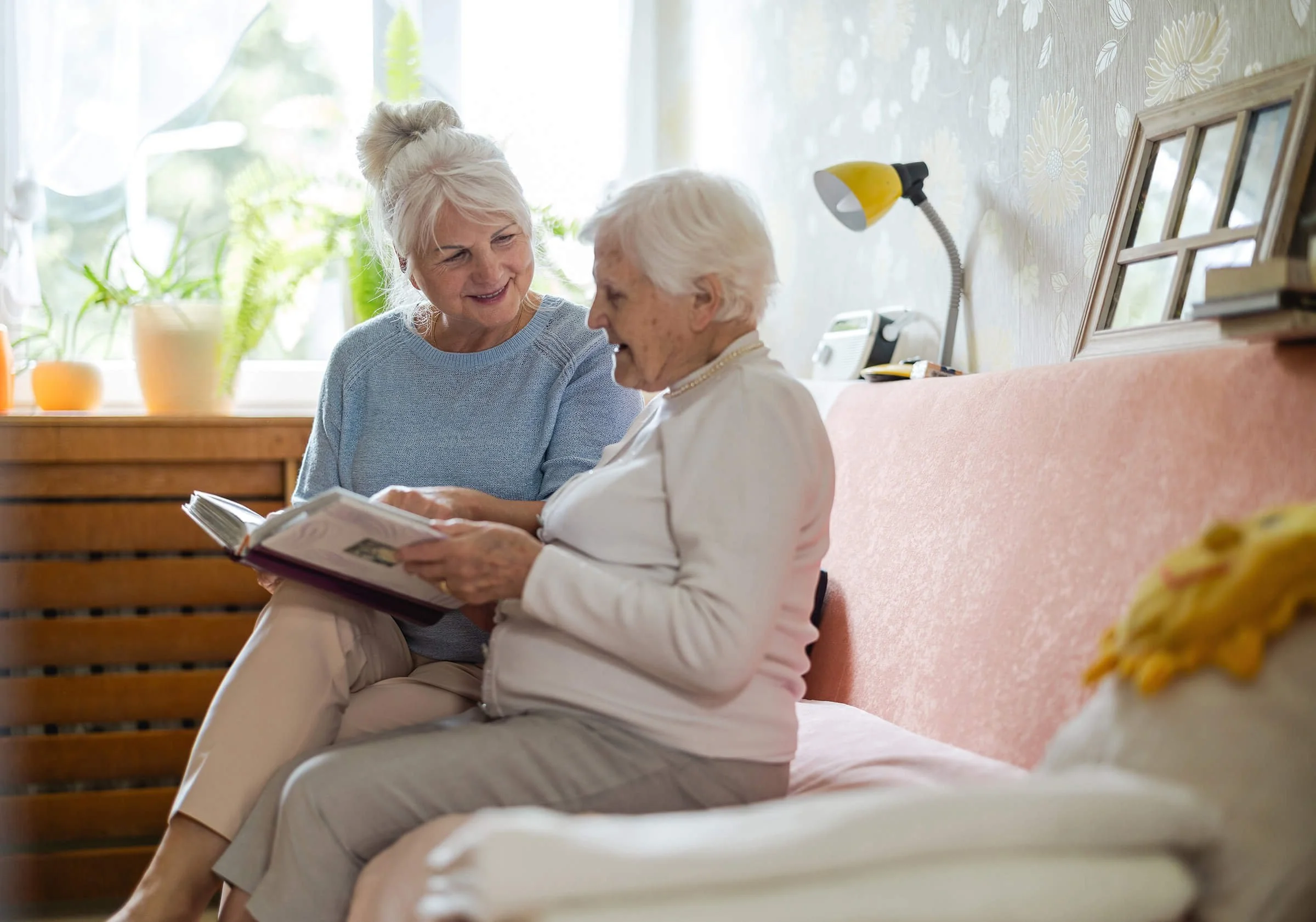 Two older women sitting together reading a book, showing companionship and comfort through senior living support.
