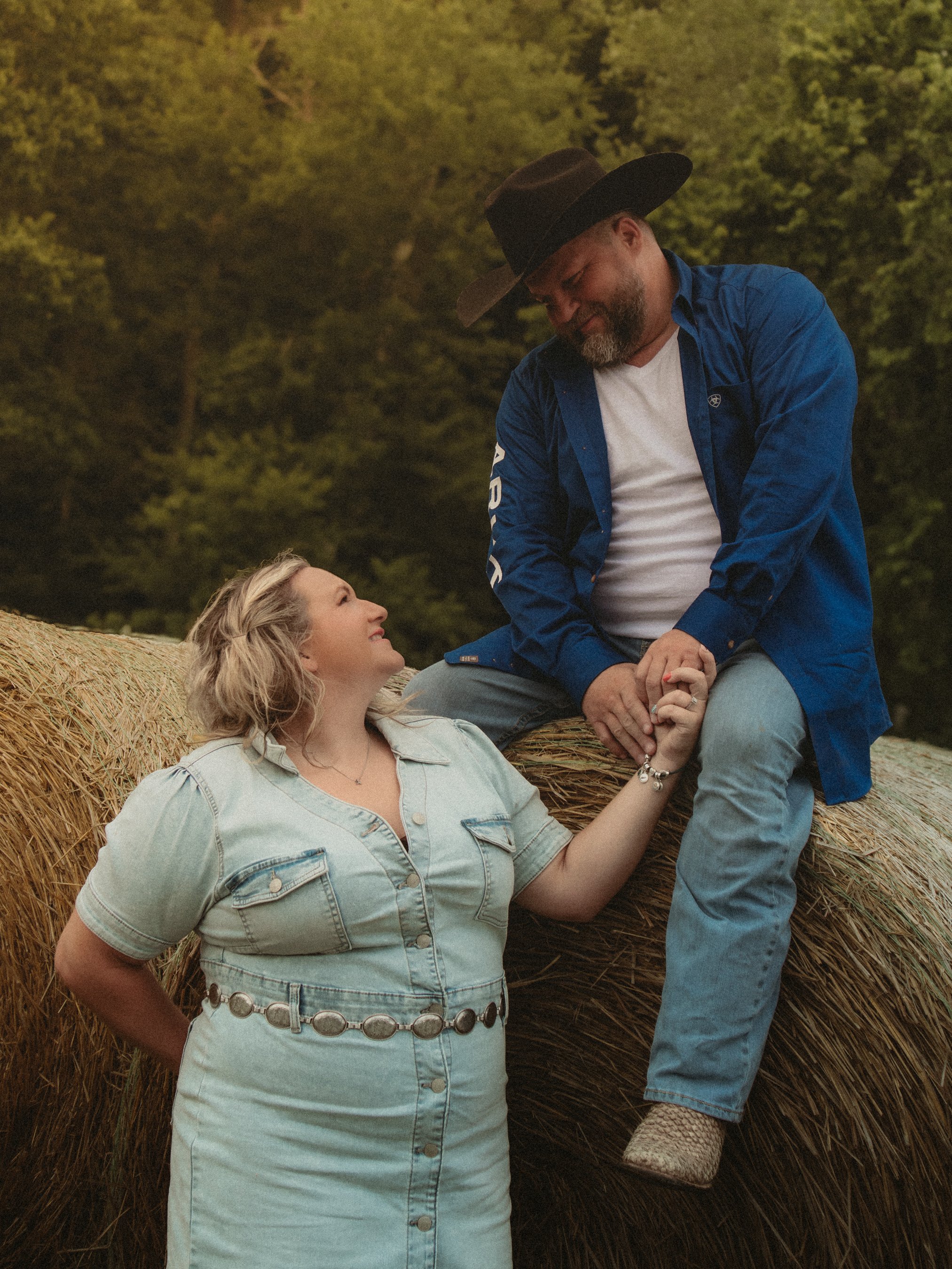 A man in a black cowboy hat, blue jacket, and white t-shirt sitting on a hay bale, holding hands with a woman in a light denim dress standing beside him, both smiling and looking at each other in a rural outdoor setting.