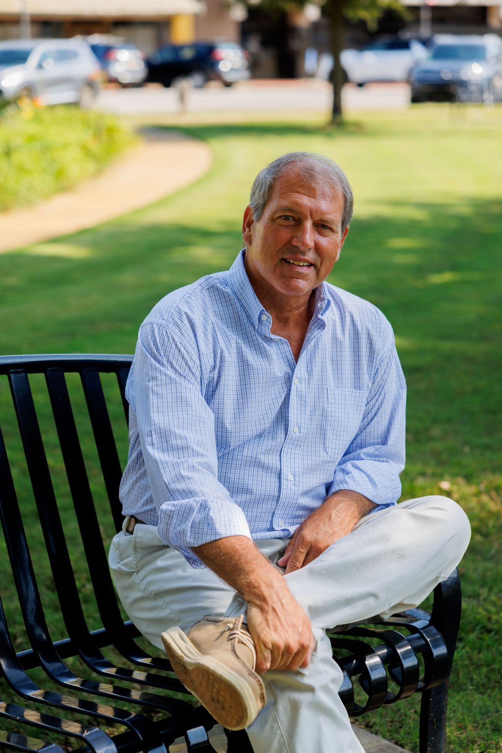 Jeffrey W. Massey sitting on a black metal park bench, wearing a blue checkered shirt and beige pants.