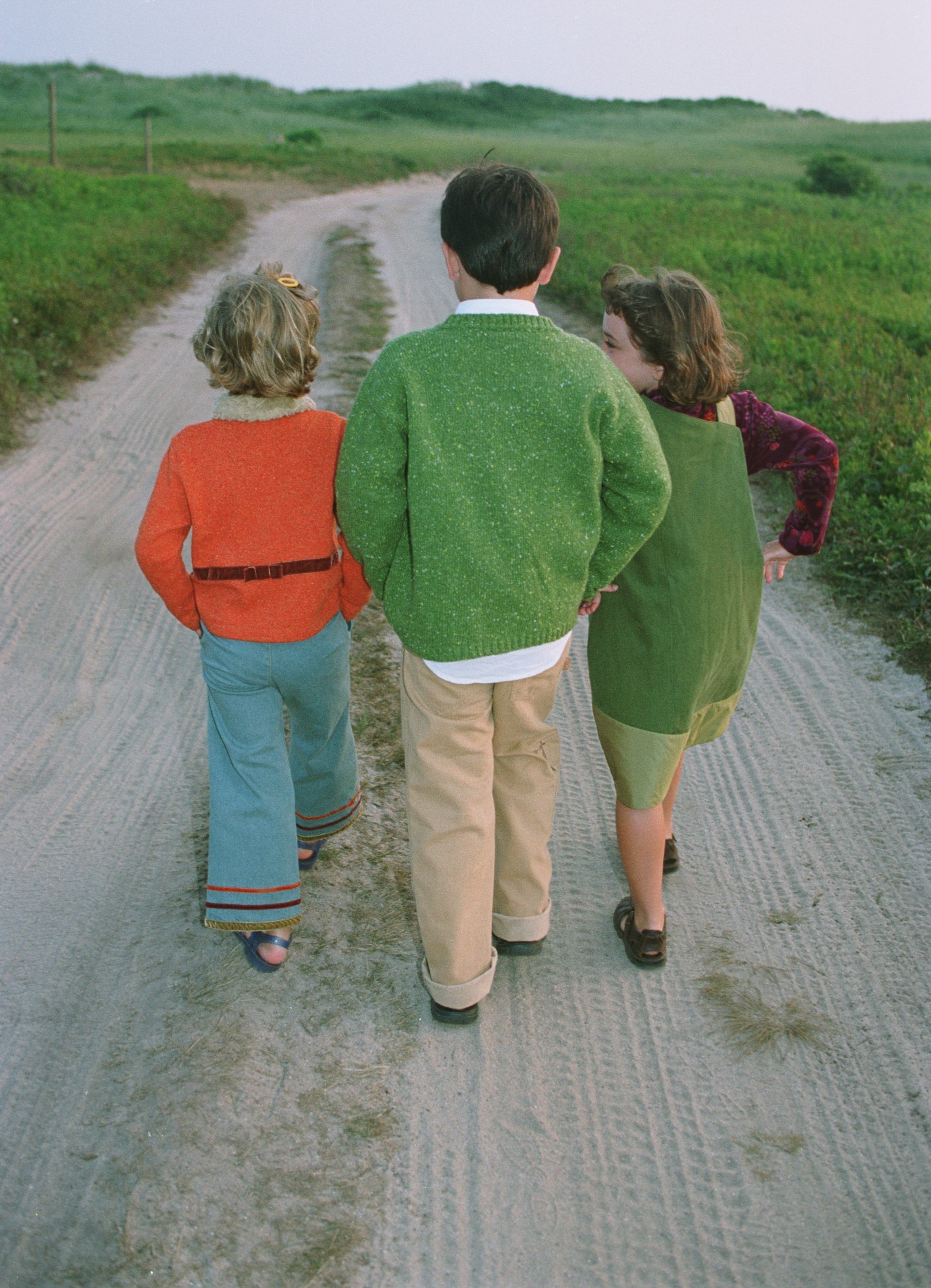 Three children walking on a dirt path in casual clothes, one in an orange sweater and blue pants, another in a green sweater and khaki pants, and the third in a green dress with a purple shirt. The path is surrounded by grass.