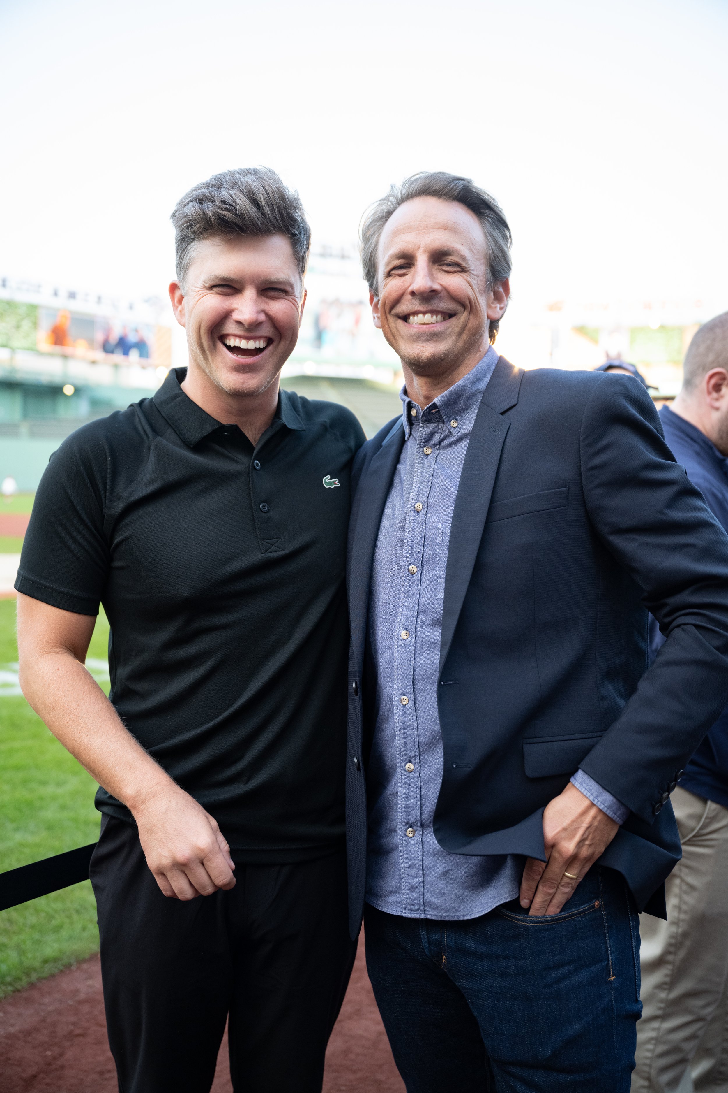 Two smiling men standing at a baseball field, one in a black polo shirt and the other in a blue jacket and shirt.