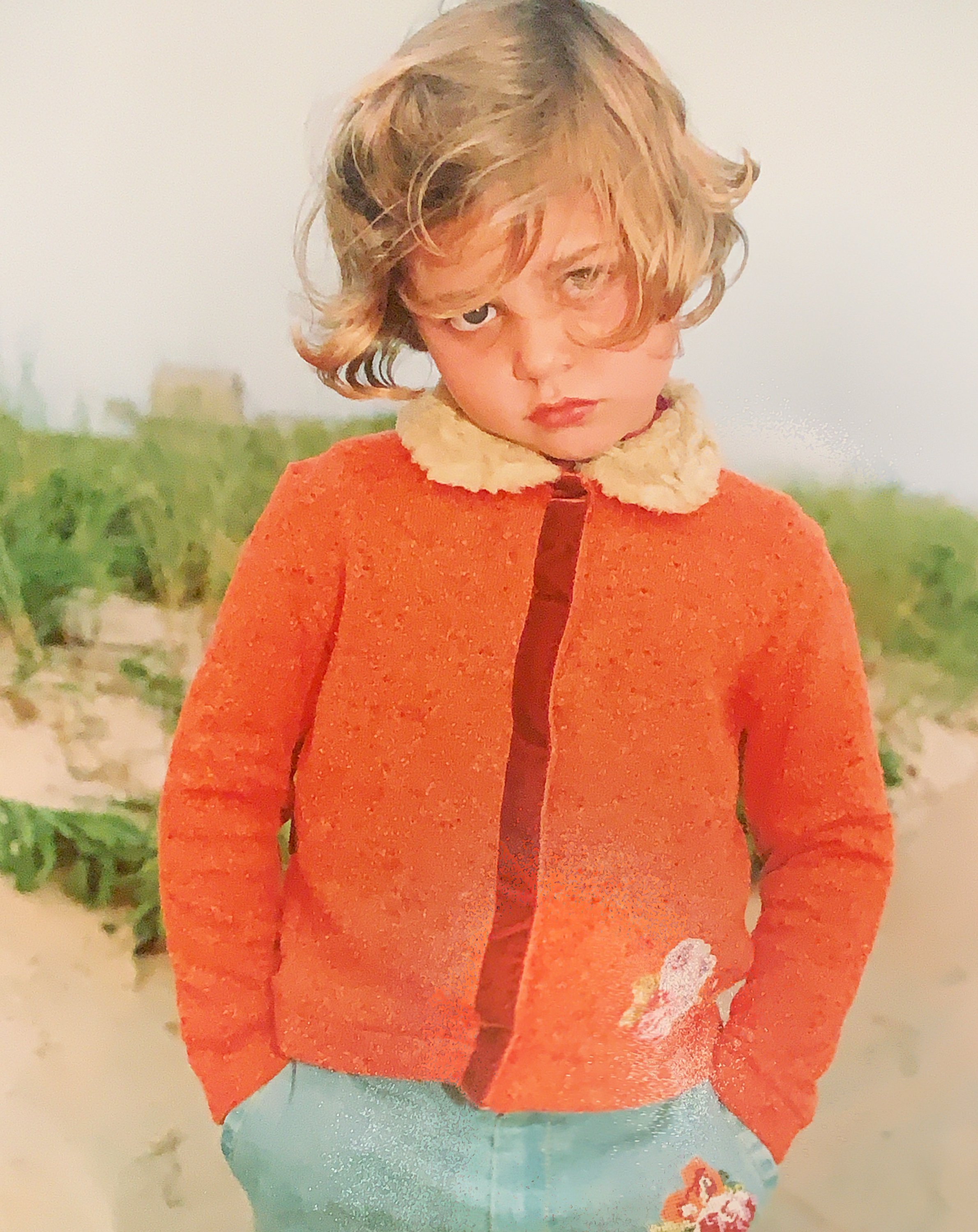 Child with curly blonde hair wearing an orange sweater and standing on a sandy beach with greenery in the background.