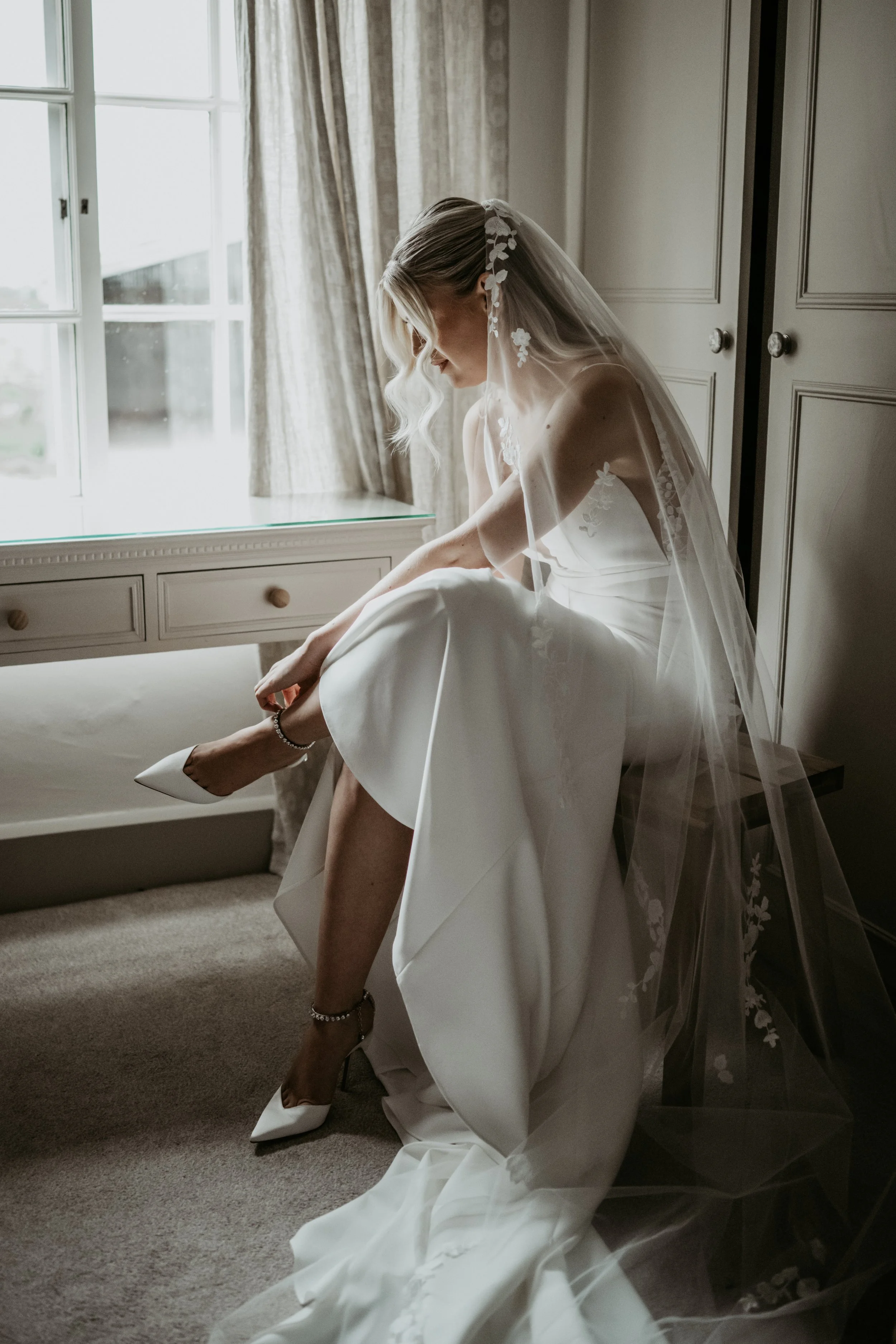 Bride in wedding dress and veil sitting by window, adjusting shoe.